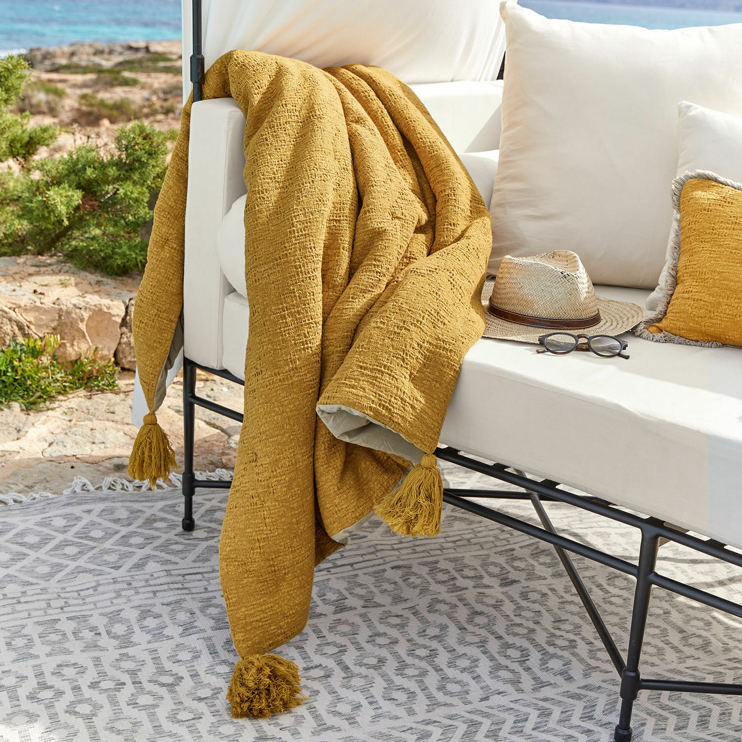 Plaid Rabea Gelbe Tagesdecke mit Quasten auf weissem Outdoor-Sofa, Terrasse mit Sonnenbrille und Hut dekoriert