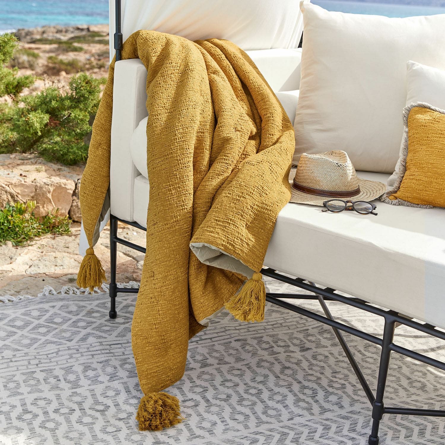 Plaid Rabea Gelbe Tagesdecke mit Quasten auf weissem Outdoor-Sofa, Terrasse mit Sonnenbrille und Hut dekoriert