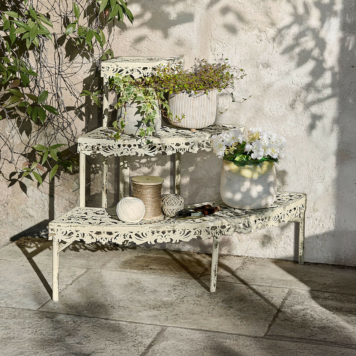 Cremefarbene Metall-Etagere mit filigranem Muster, bepflanzt und mit Gartengeräten auf Steinboden