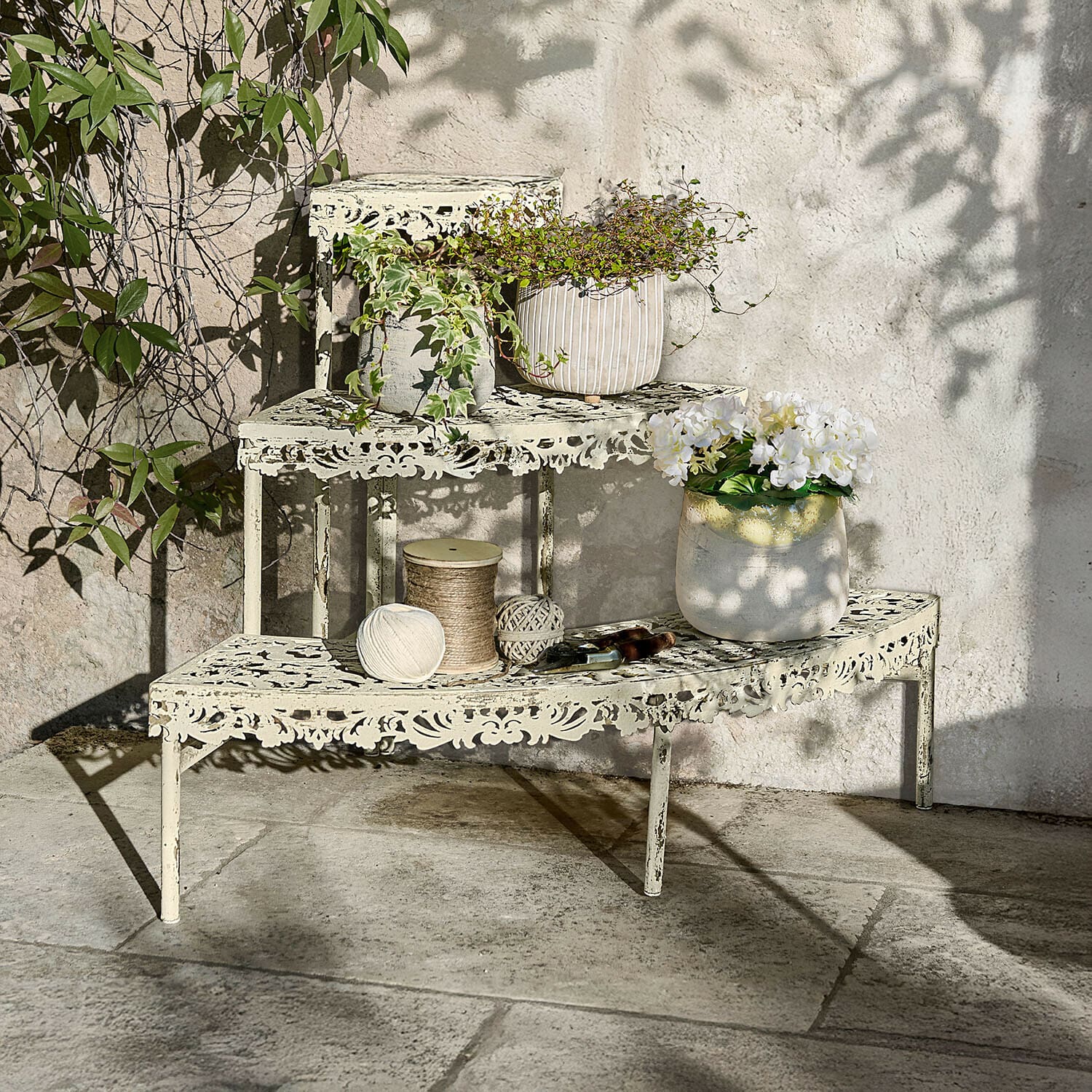 Pflanztreppe Cresselay Cremefarbene Metall-Etagere mit filigranem Muster, bepflanzt und mit Gartengeräten auf Steinboden