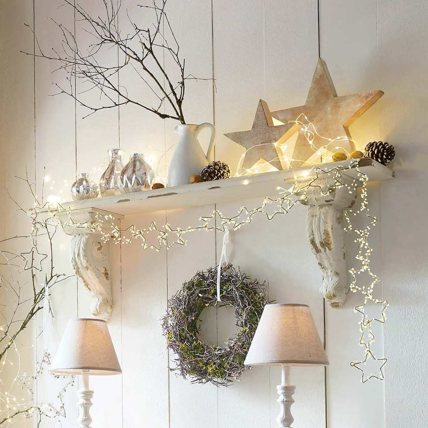 Weisses Holzregal mit sternförmiger Lichterkette, Kranz und Tischlampen im Landhausstil