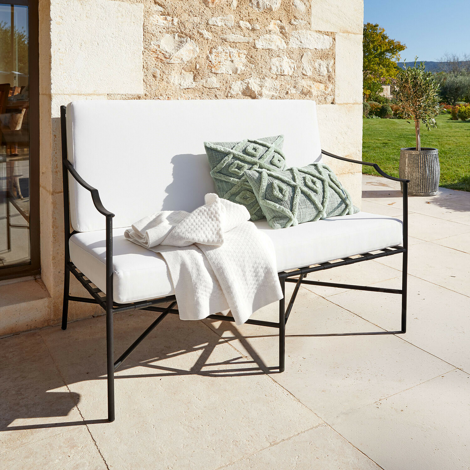 Sofa Vexford Weisse Metall-Gartenbank mit weichen Polstern und grünen Dekokissen auf sonniger Terrasse