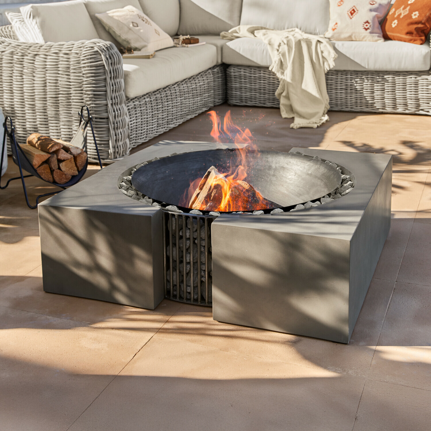 Feuerstelle Charonville Moderne Feuerstelle aus schwarzem Metall mit Steinen auf Terrasse neben Rattansofa mit Kissen