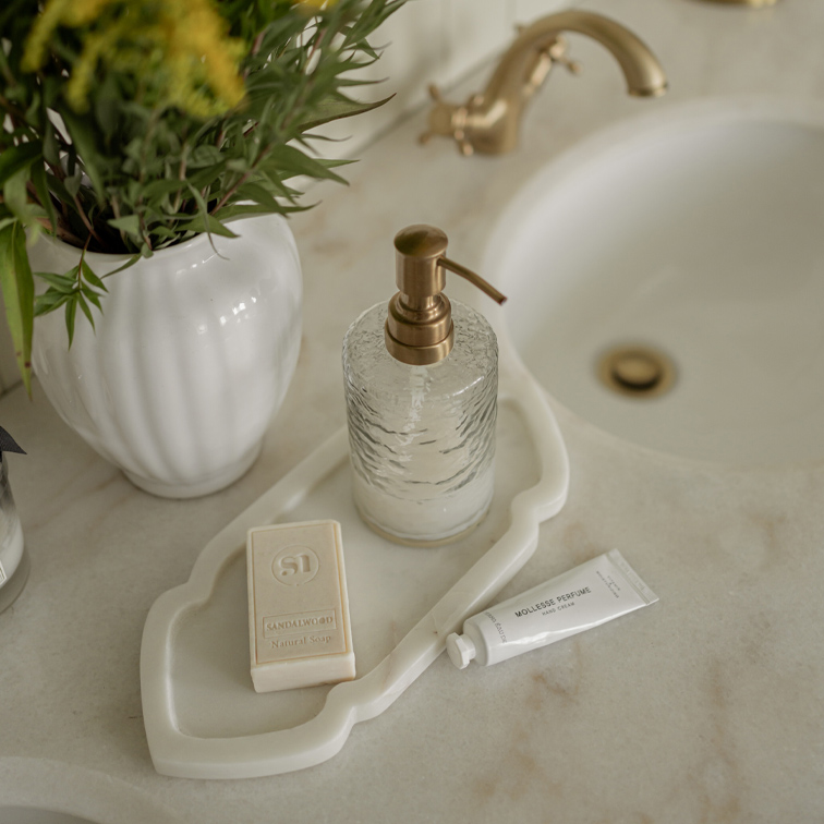 Glas-Seifenspender mit Messingpumpe auf weissem Tablett neben Seife und Handcreme auf Marmorwaschbecken