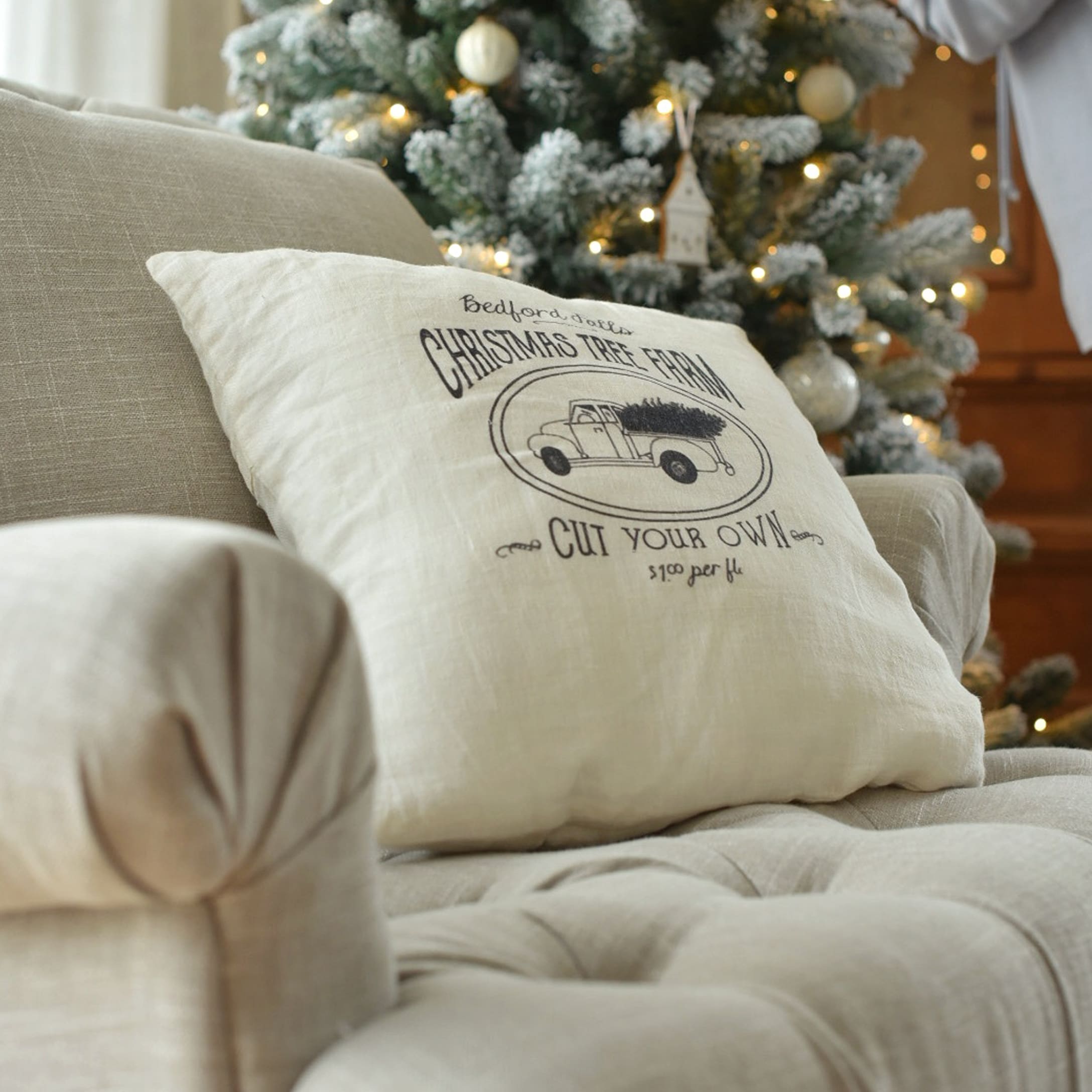 Beiges Leinenkissen mit Weihnachtsauto-Motiv auf Sofa vor geschmücktem Tannenbaum mit Lichtern