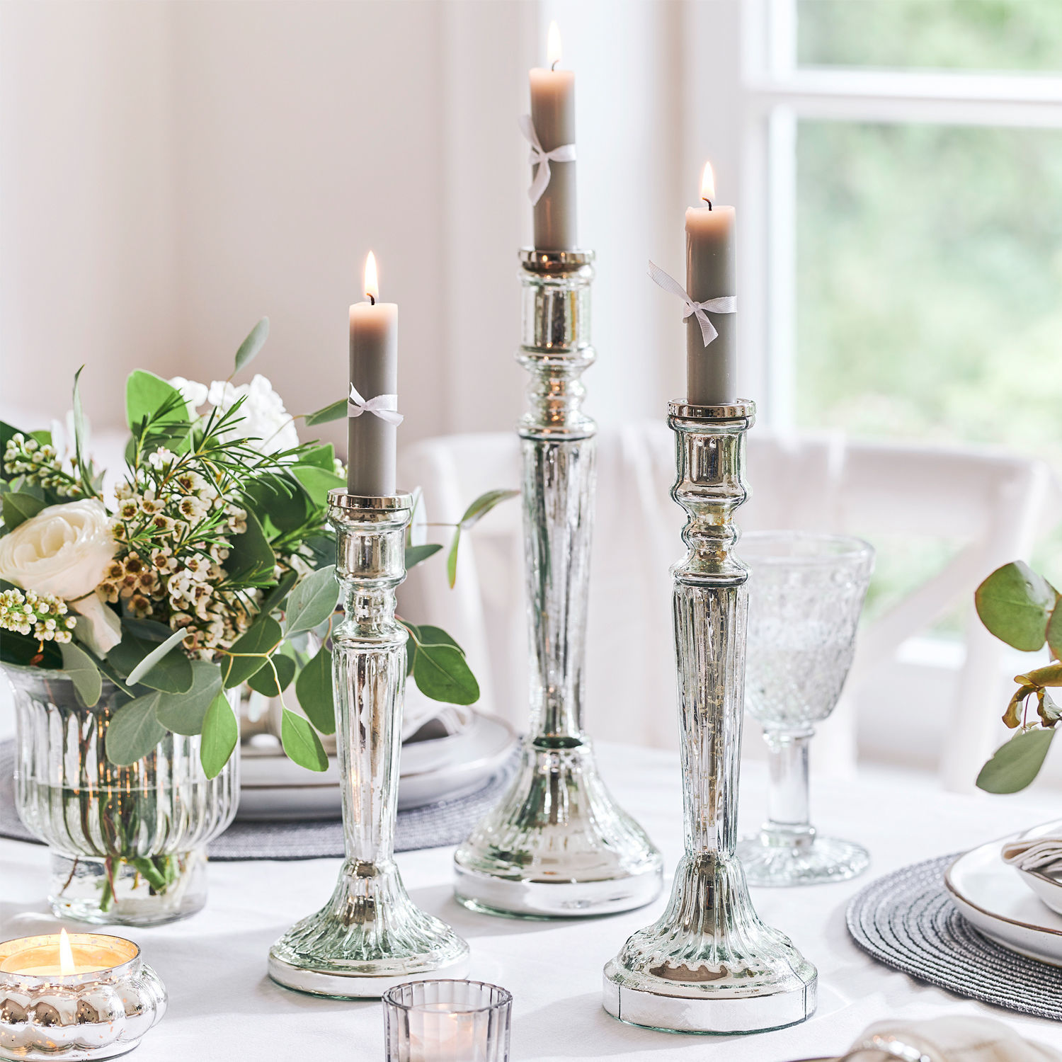 Kerzenständer 3er Set Riley Drei silberne Kerzenhalter mit grauen Kerzen auf weiss gedecktem Tisch mit Blumen und Glas