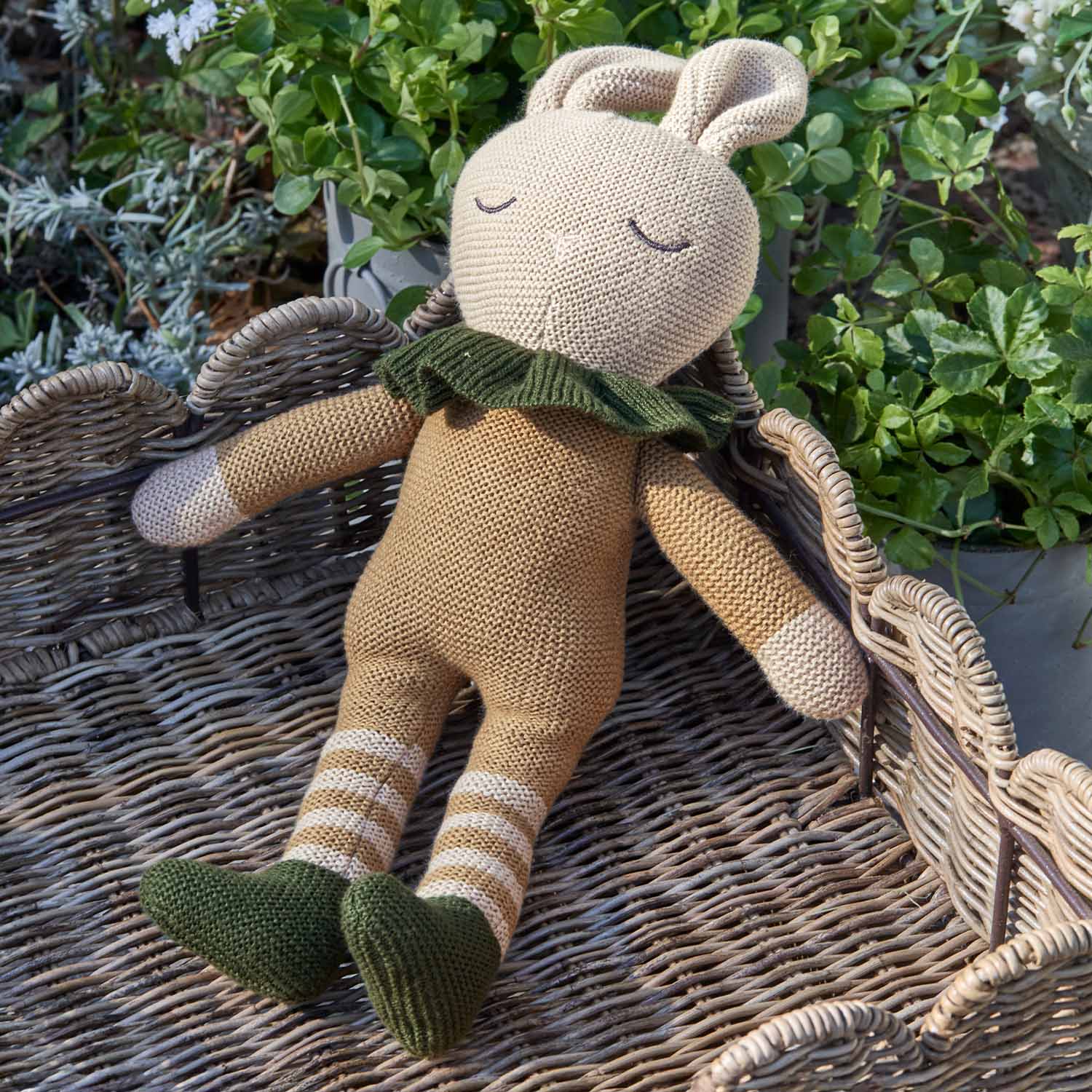 Beige gestricktes Hasenstofftier mit grünen Schuhen und Kragen auf Korbgeflecht, Kinderspielzeug