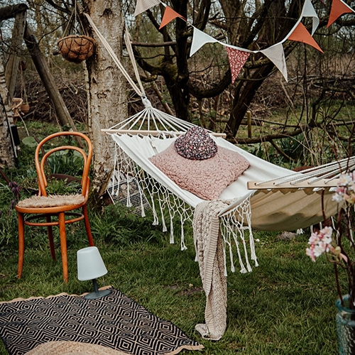 Weisse Stoffhängematte mit rosa Kissen und Decke auf grünem Rasen, Holzstuhl und Girlande im Garten