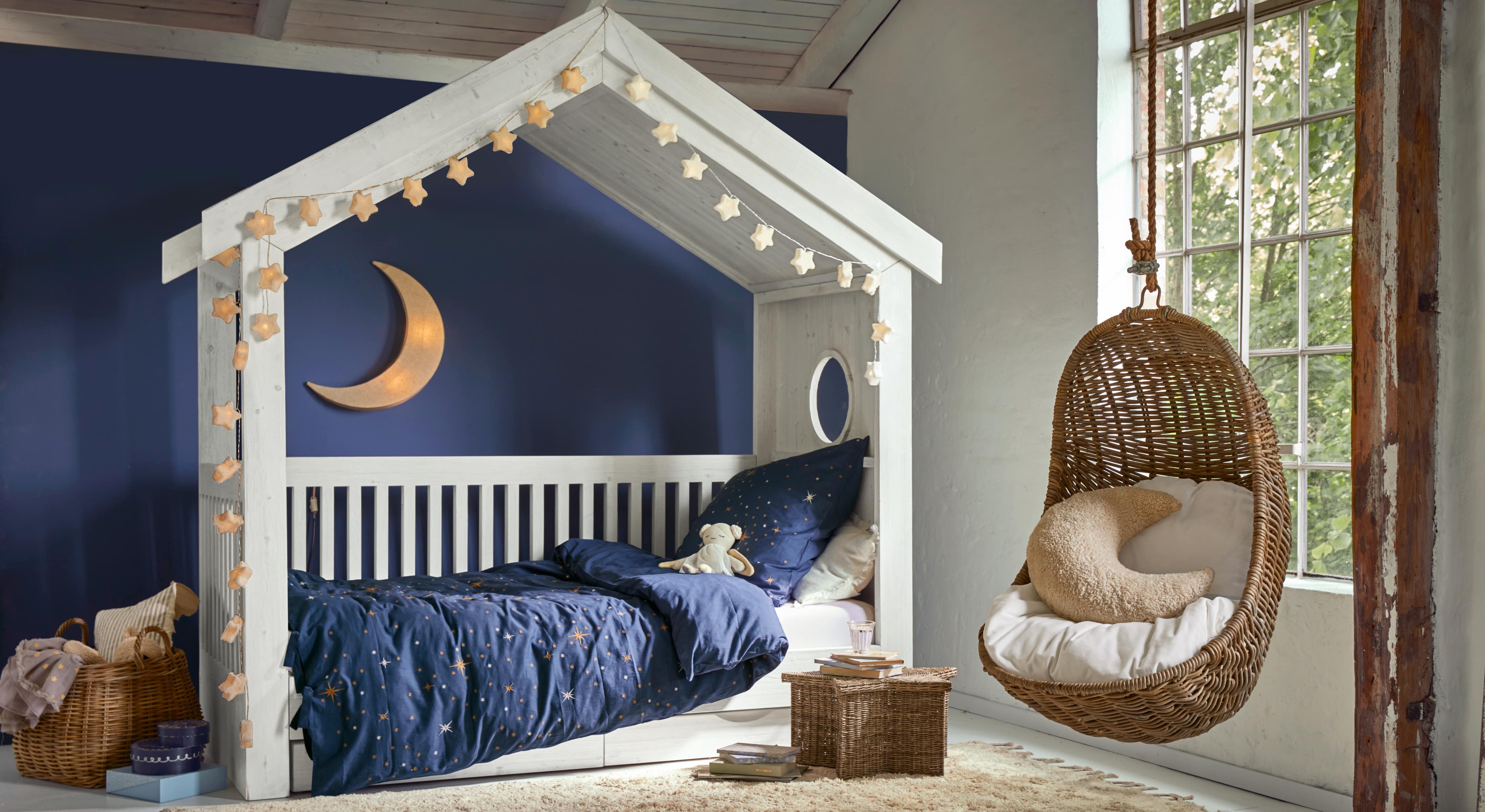 Weisses Holzhaus-Bett mit dunkelblauer Sternenbettwäsche und hängendem Korbsessel im Kinderzimmer