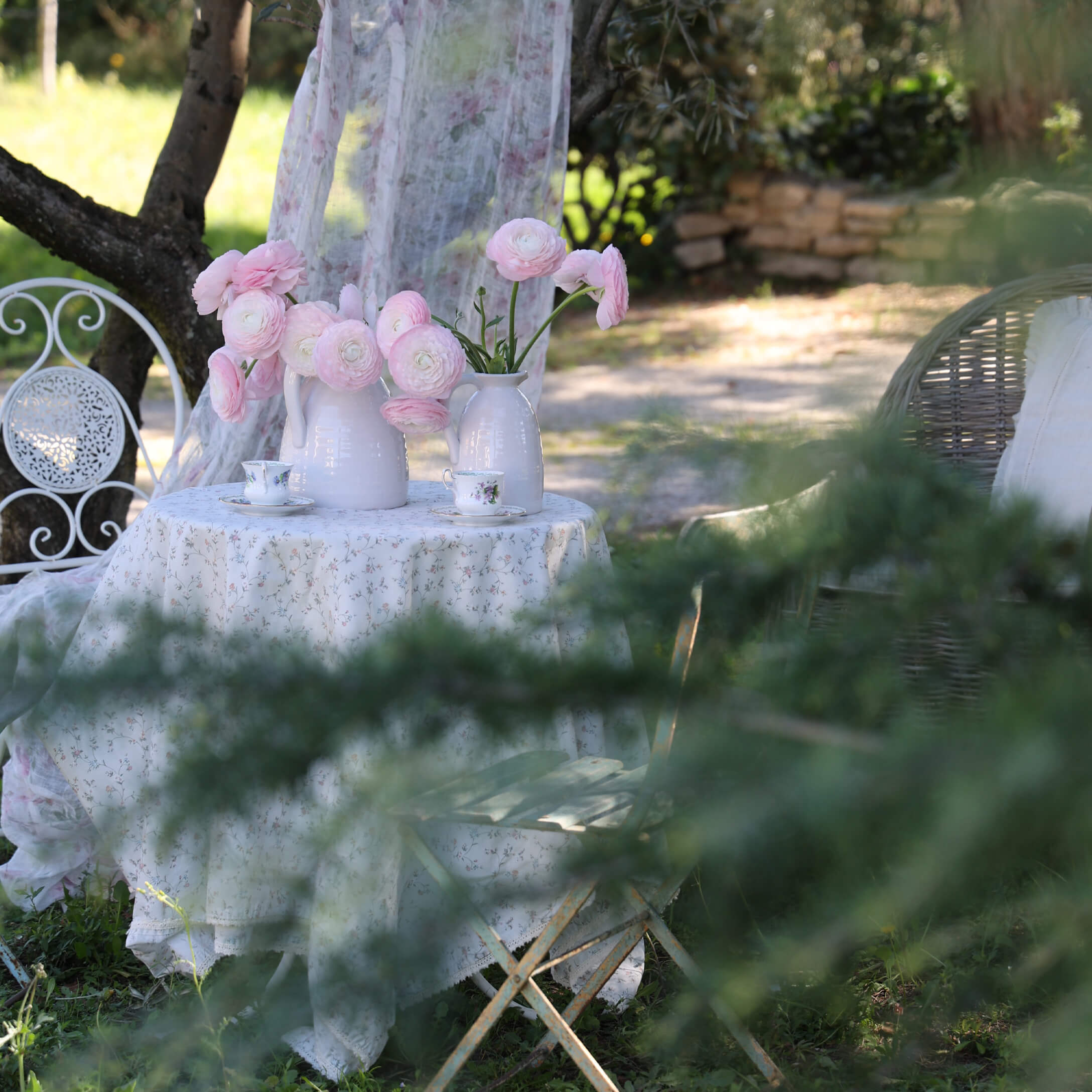 Weißer Gartentisch mit geblümter Tischdecke und weißen Krügen mit rosa Blumen, romantische Stimmung