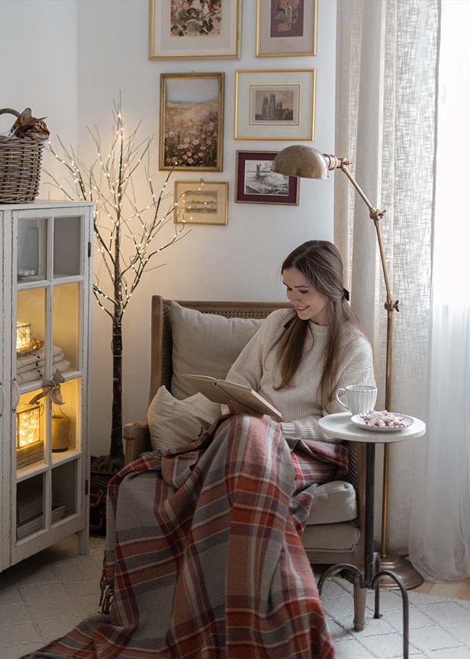 Frau liest Buch unter warmem Plaid im gemütlichen Wohnzimmer mit Stehleuchte und Wandbildern.