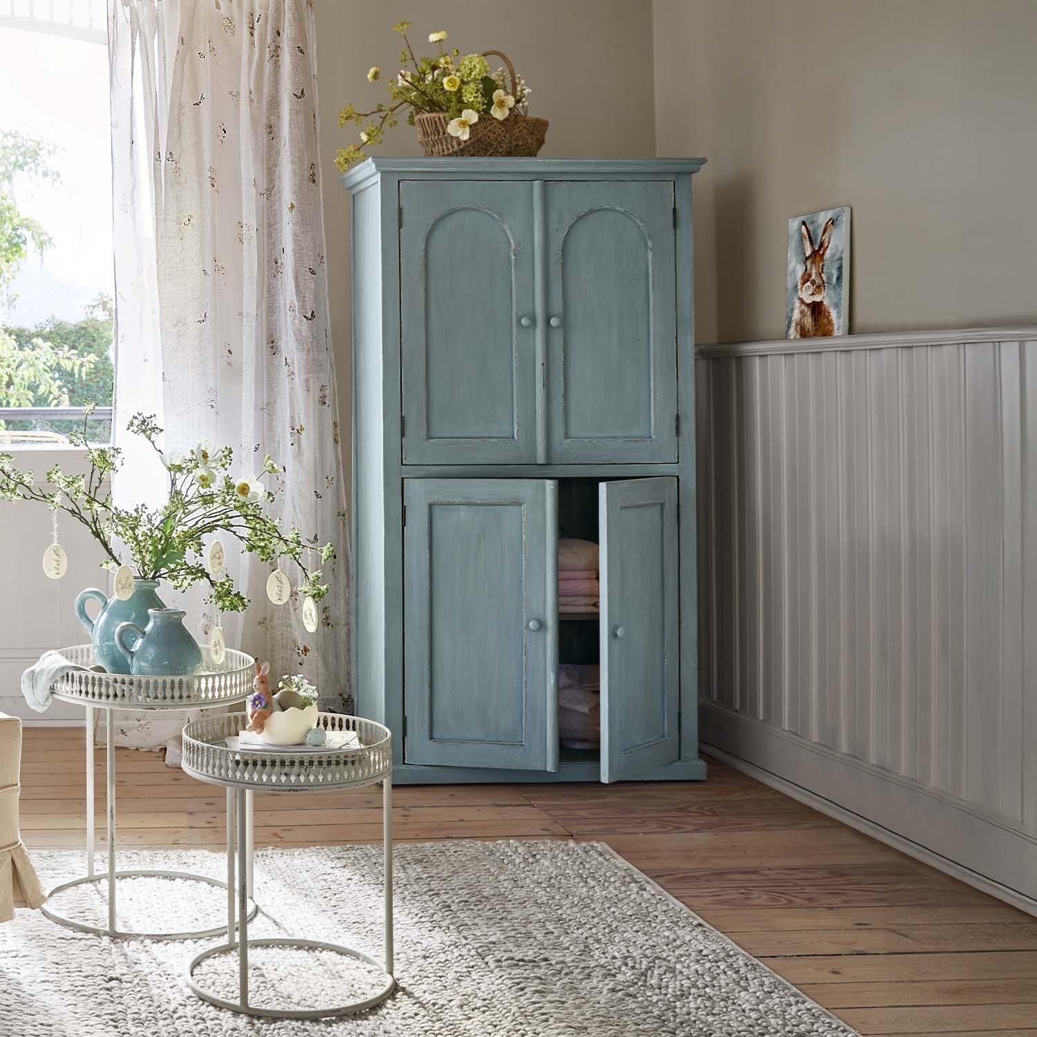 Hellblauer Holzschrank mit zwei Türen in gemütlichem Raum mit Blumenarrangements und Teppich