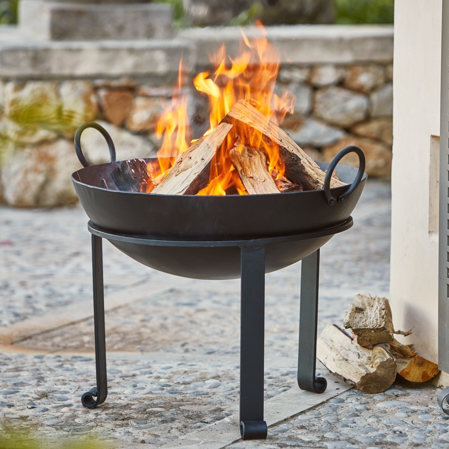 Feuerschale Harrogate Schwarzer Metall-Feuerkorb mit brennendem Holz auf steiniger Terrasse im Garten