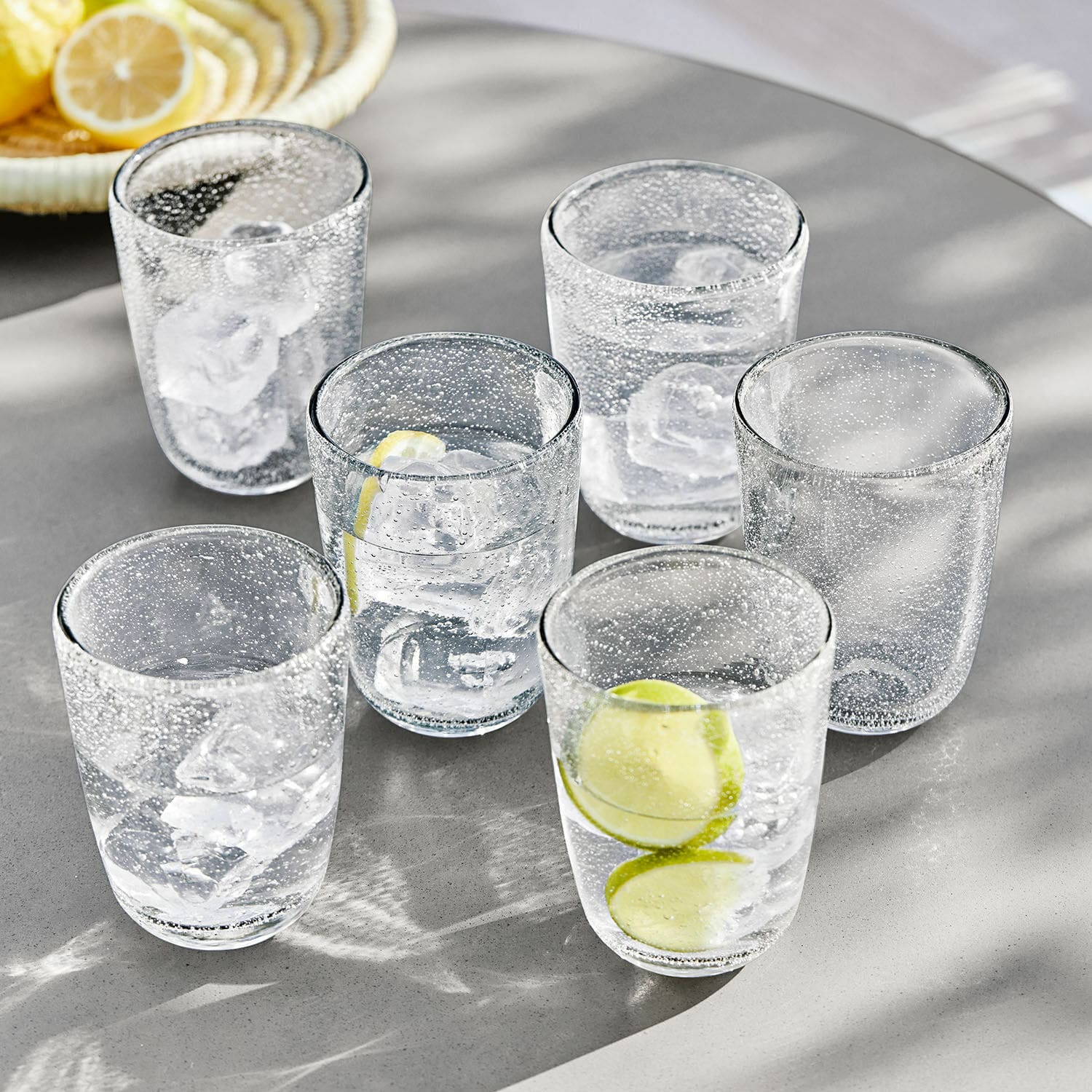Sechsteiliges Set Gläser aus klarem Glas mit Luftblasen, Eiswürfeln und Zitronenscheiben auf grauem Tisch
