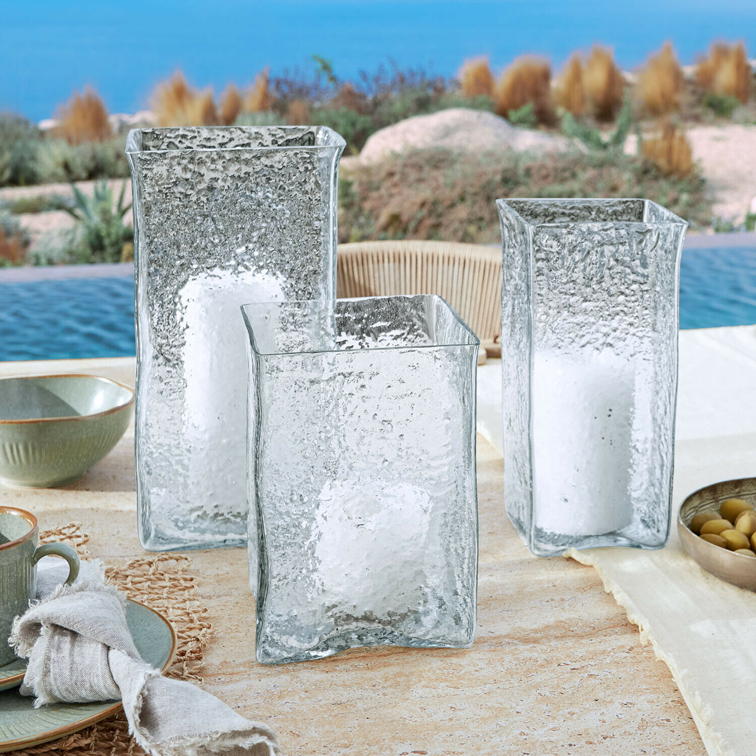 Windlicht 3er Set Xarlac Drei durchsichtige, strukturierte Glaslaternen mit weissen Kerzen auf einem Tisch beim Pool