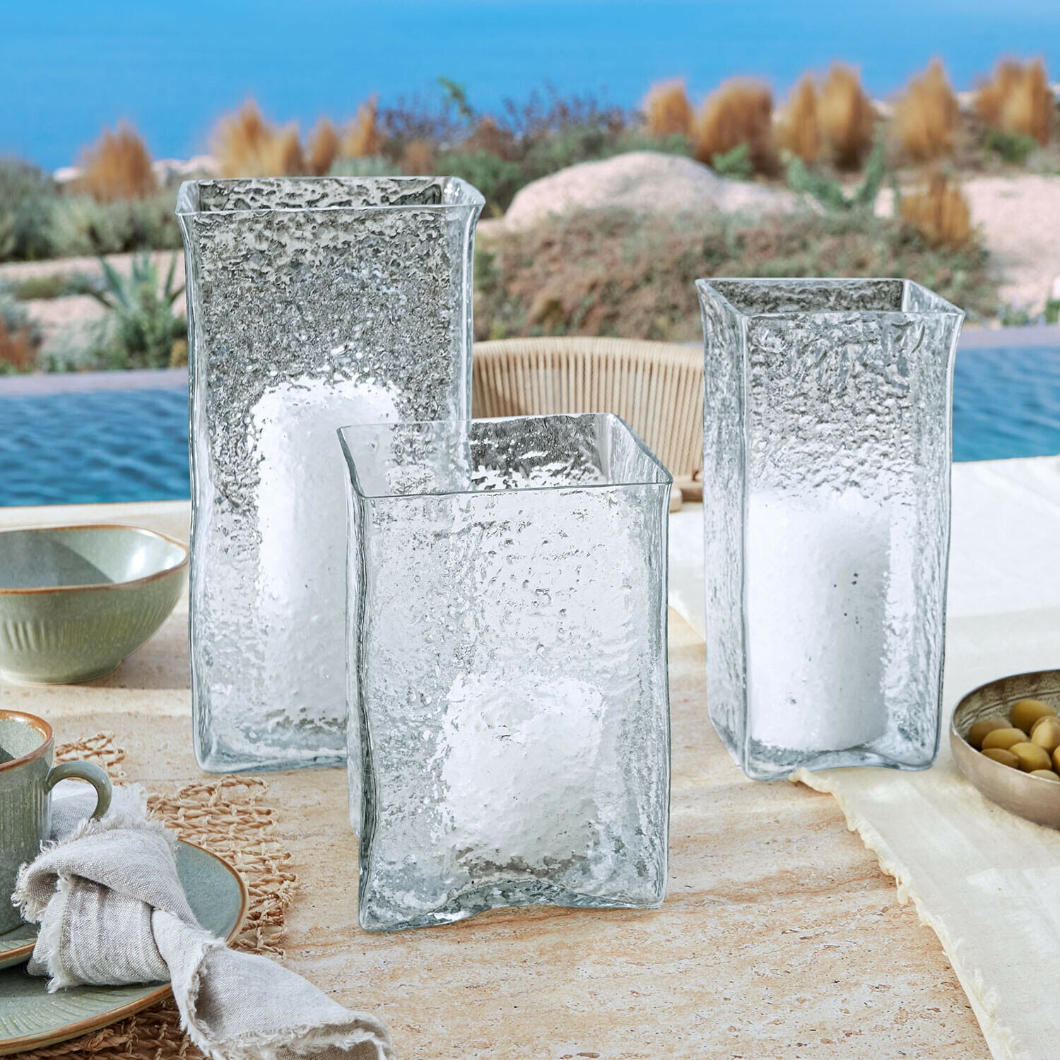 Windlicht 3er Set Xarlac Drei durchsichtige, strukturierte Glaslaternen mit weissen Kerzen auf einem Tisch beim Pool