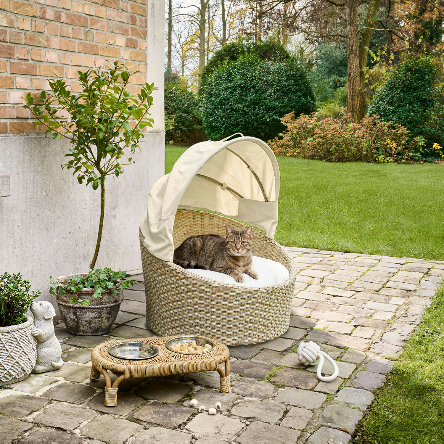 In- & Outdoor-Haustierkorb Brinelza Ovales Katzenbett aus beigem Rattan mit weissem Kissen und Dach auf gepflasterter Terrasse im Garten