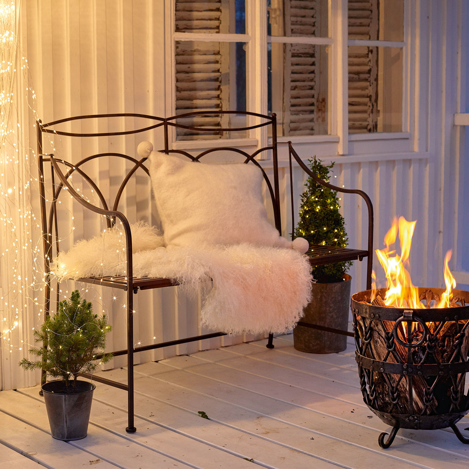 Bank Jablines Metallbank mit weissem Kissen und Fellüberwurf neben Feuerkorb auf Terrasse mit Lichterketten-Dekor