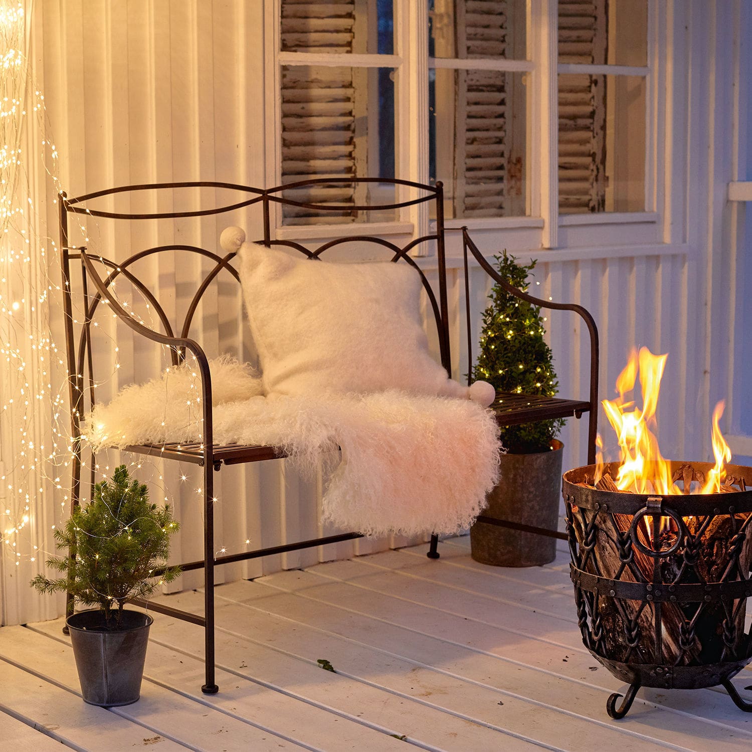 Bank Jablines Metallbank mit weissem Kissen und Fellüberwurf neben Feuerkorb auf Terrasse mit Lichterketten-Dekor