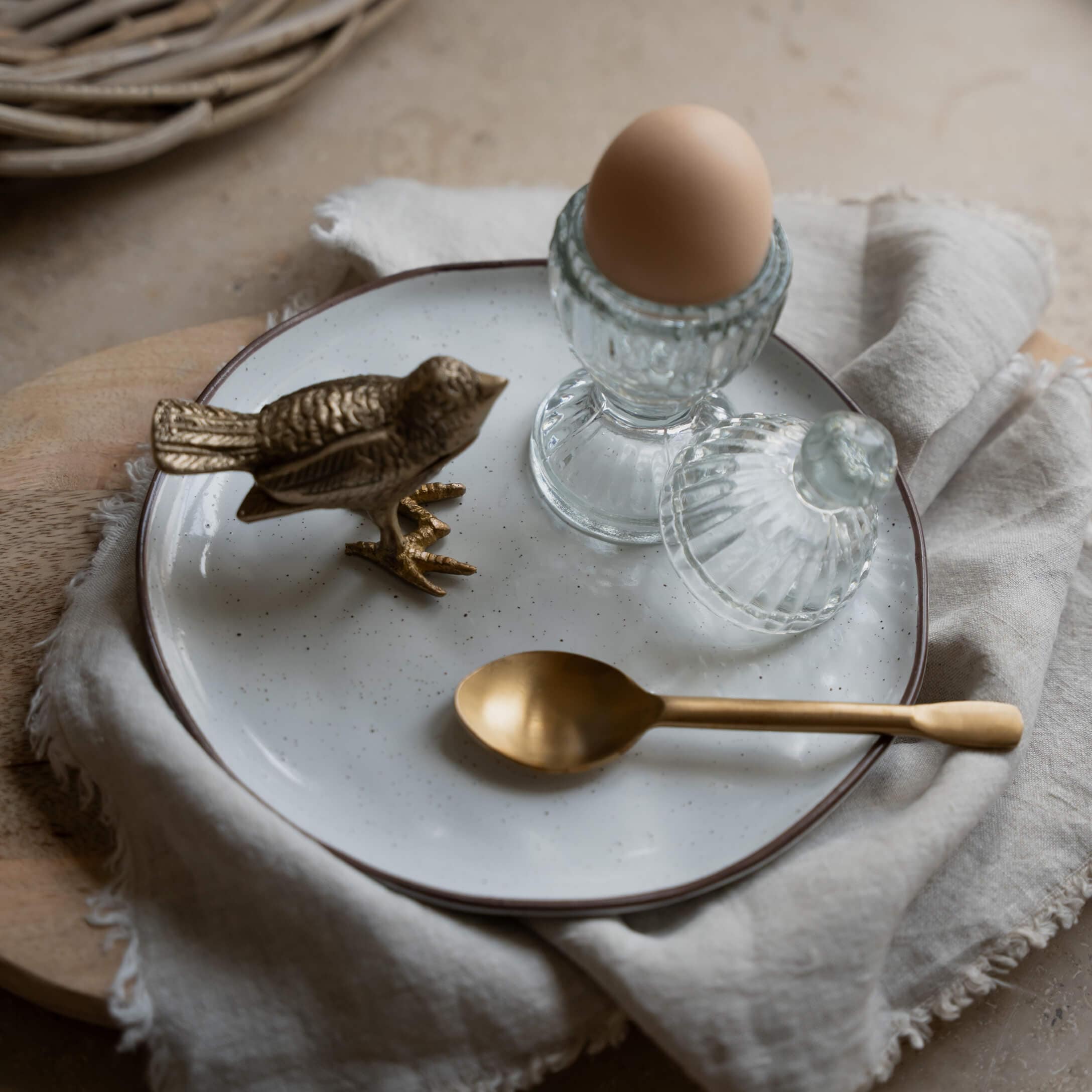 Glas-Eierbecher mit Deckel und braunem Ei auf weissem Teller mit Messinglöffel und Vogel-Figur