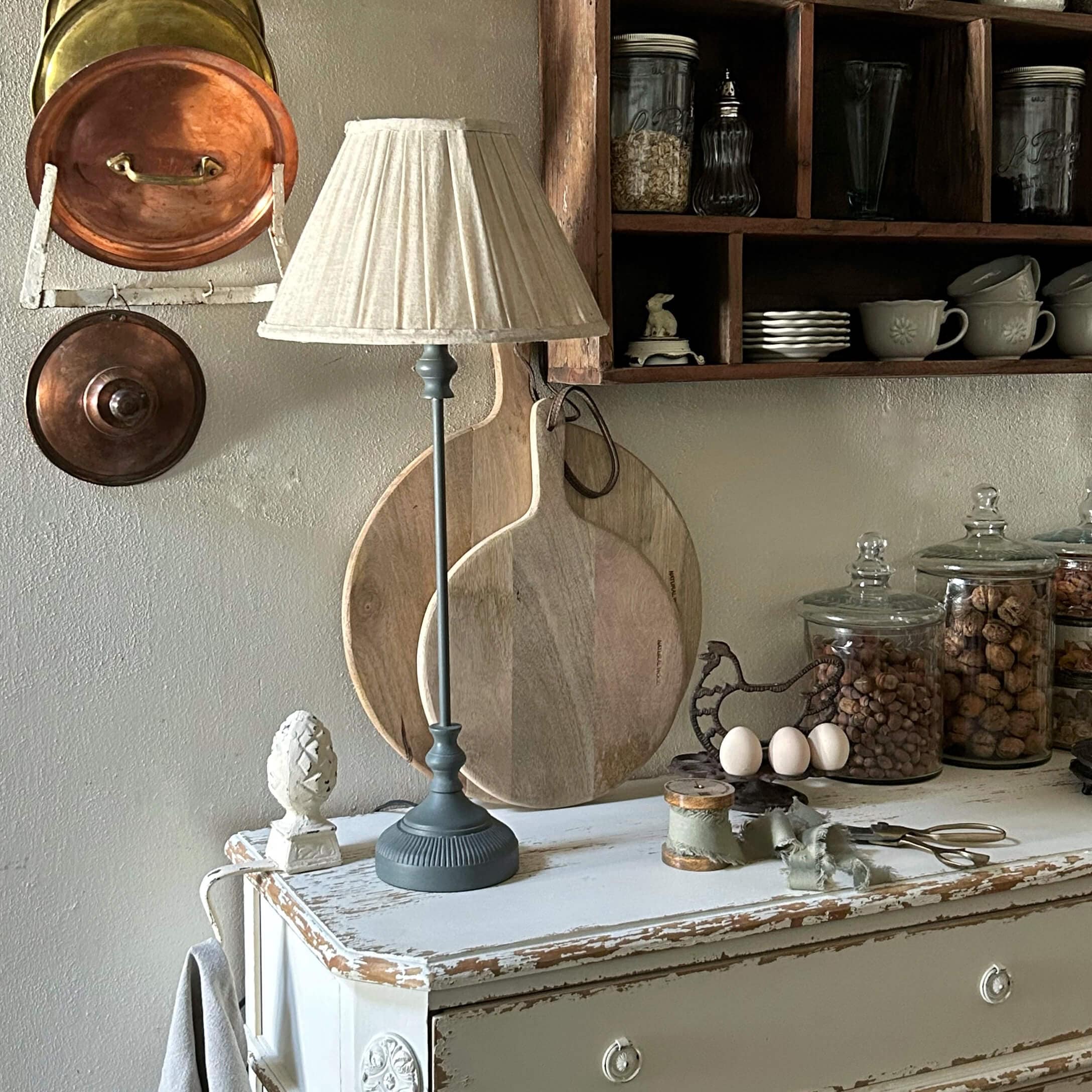 Tischlampe mit grauem Sockel und cremefarbenem Schirm auf weissem, antik wirkendem Schrank in rustikaler Küche
