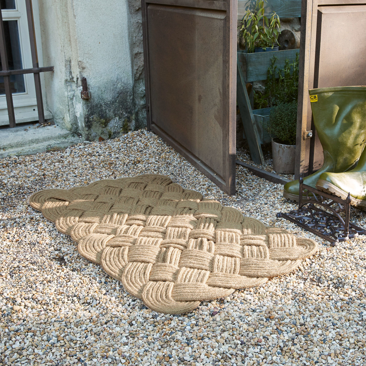 Beige Fussmatte aus geflochtenem Seil auf Kies vor offener Tür neben grünen Gummistiefeln