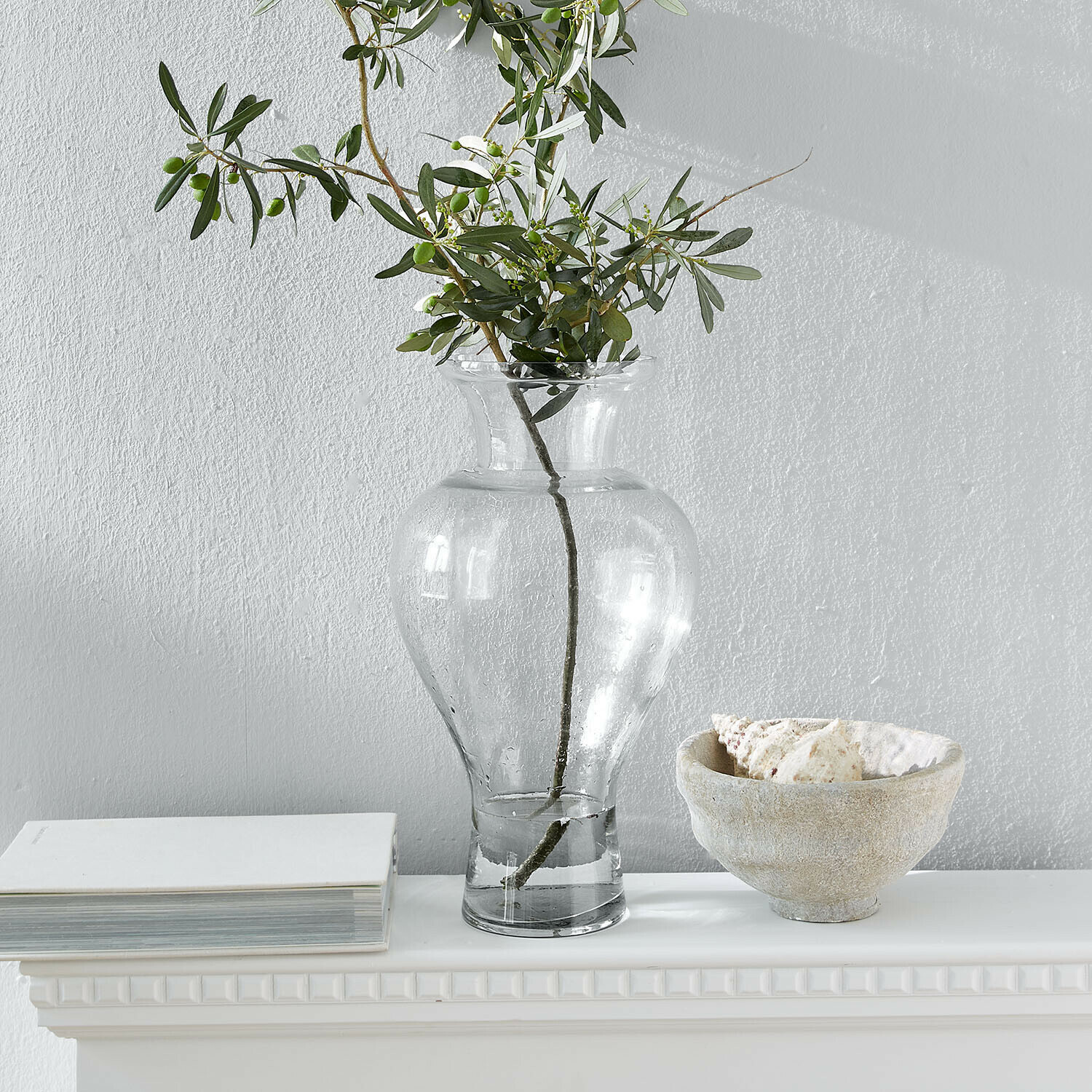 Transparente Glasvase mit grünen Zweigen auf weissem Regal mit Buch und Muschelschale