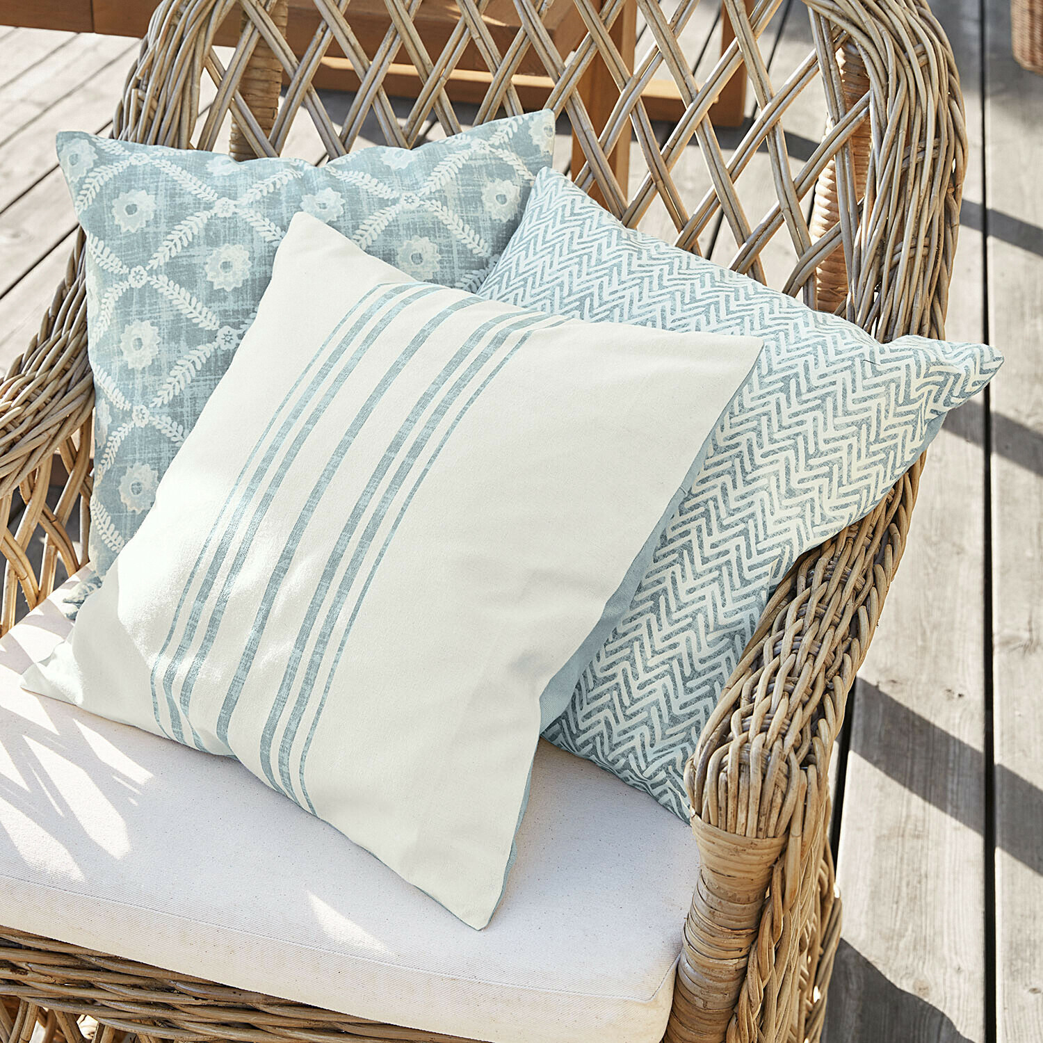 Drei dekorative Kissen in Blau- und Weiss-Tönen auf einem Rattansessel auf Holzterrasse