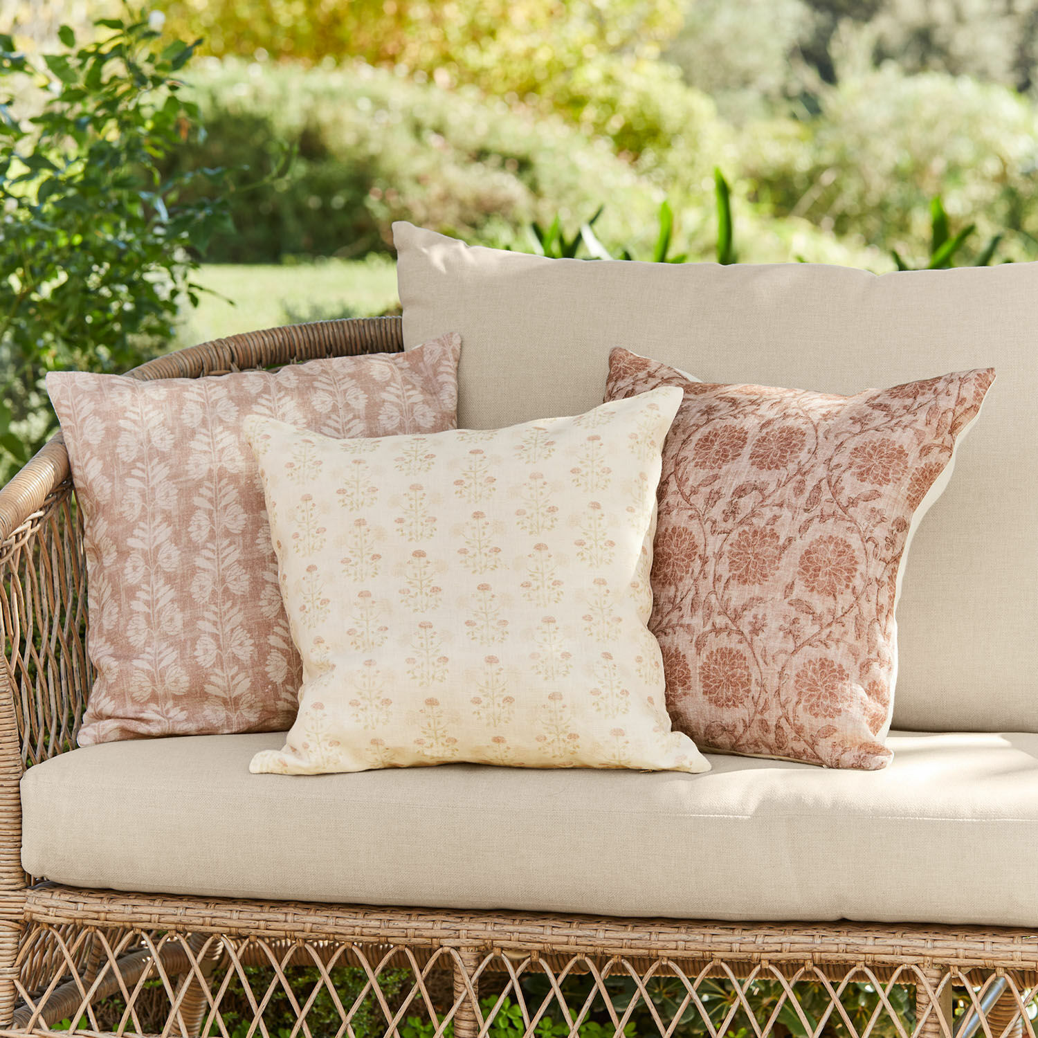 Kissenhülle 3er Set Tarouja Drei dekorative Baumwollkissen in Beige-Rosa mit floralen Mustern auf einem Rattansofa im Garten.