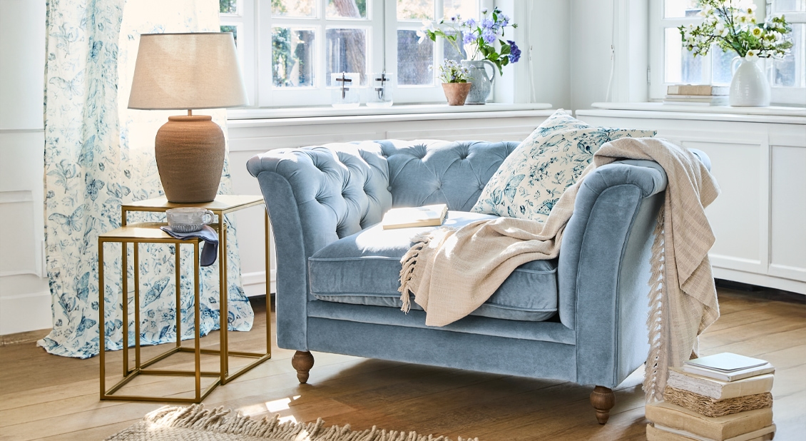 Blaues Samtsofa mit floralem Kissen und beiger Decke in hellem Wohnzimmer mit Holzboden