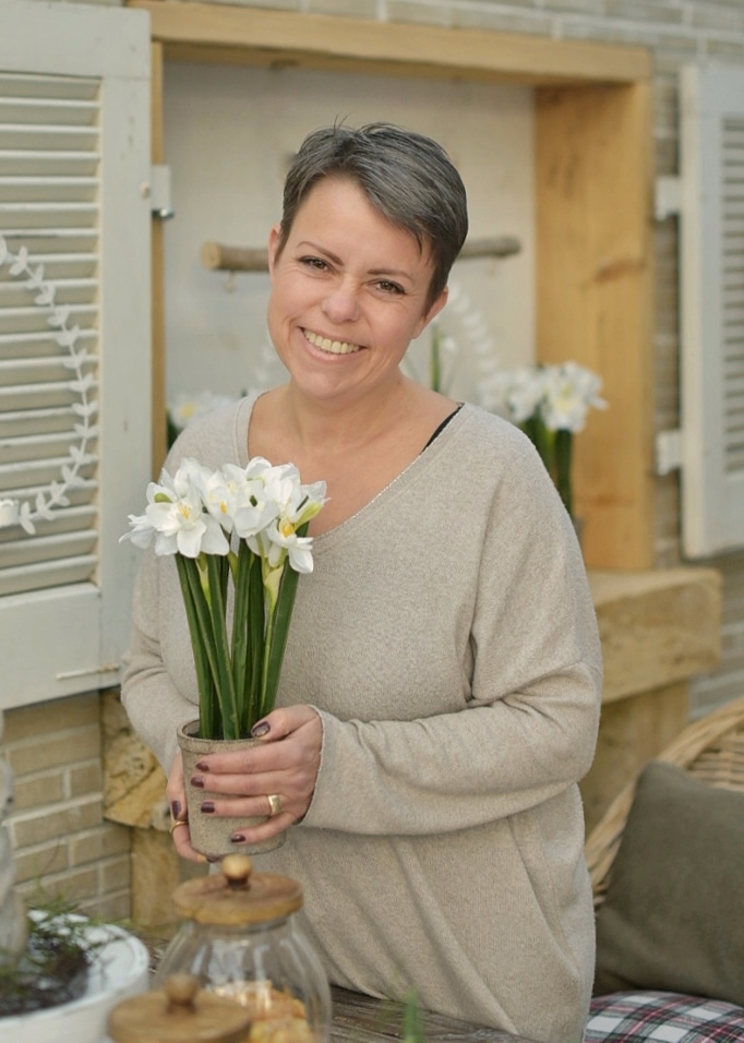 Frau hält Topf mit weissen Blumen in natürlicher Osterdekoration, trägt beigen Pullover.