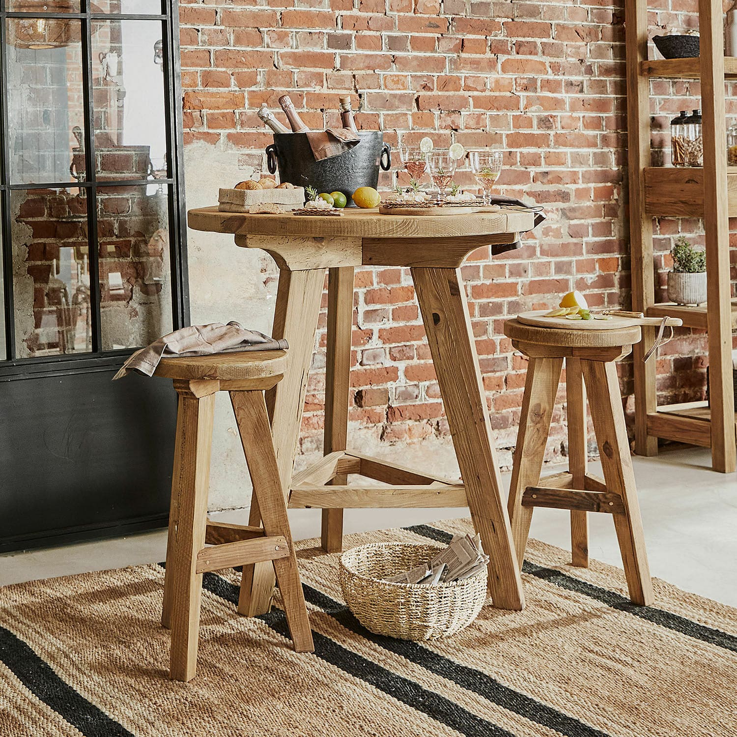 Hoher Holztisch mit zwei Hockern aus Naturholz auf Juteteppich vor Backsteinwand im Industriestil