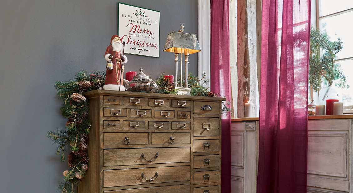Holzkommode mit vielen Schubladen, dekoriert mit Weihnachtsdekoration und Vintage-Lampe neben Fenster mit roten Vorhängen