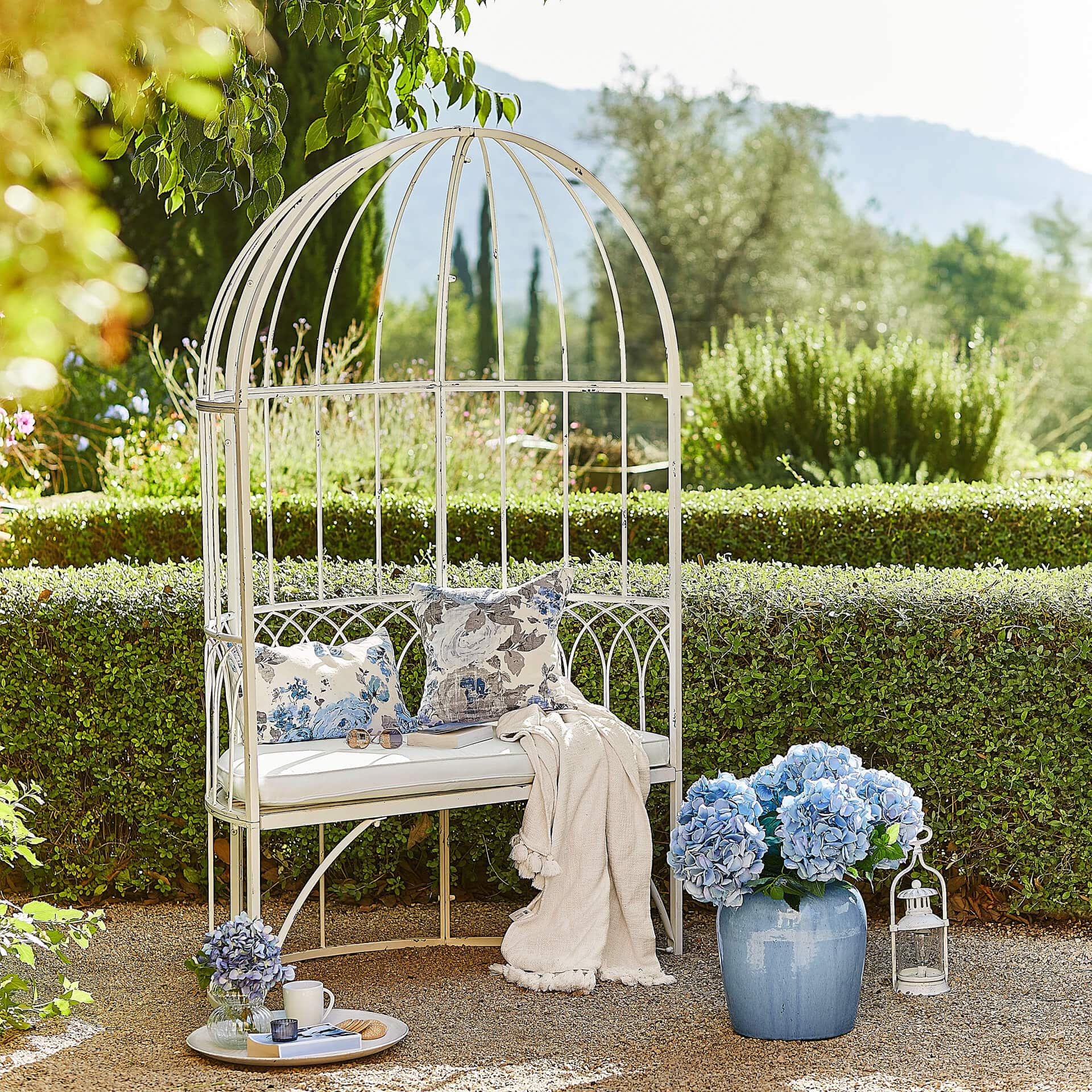 Weisse Metall-Gartenbank mit Kissen und blauen Blumen in Vase vor grüner Hecke