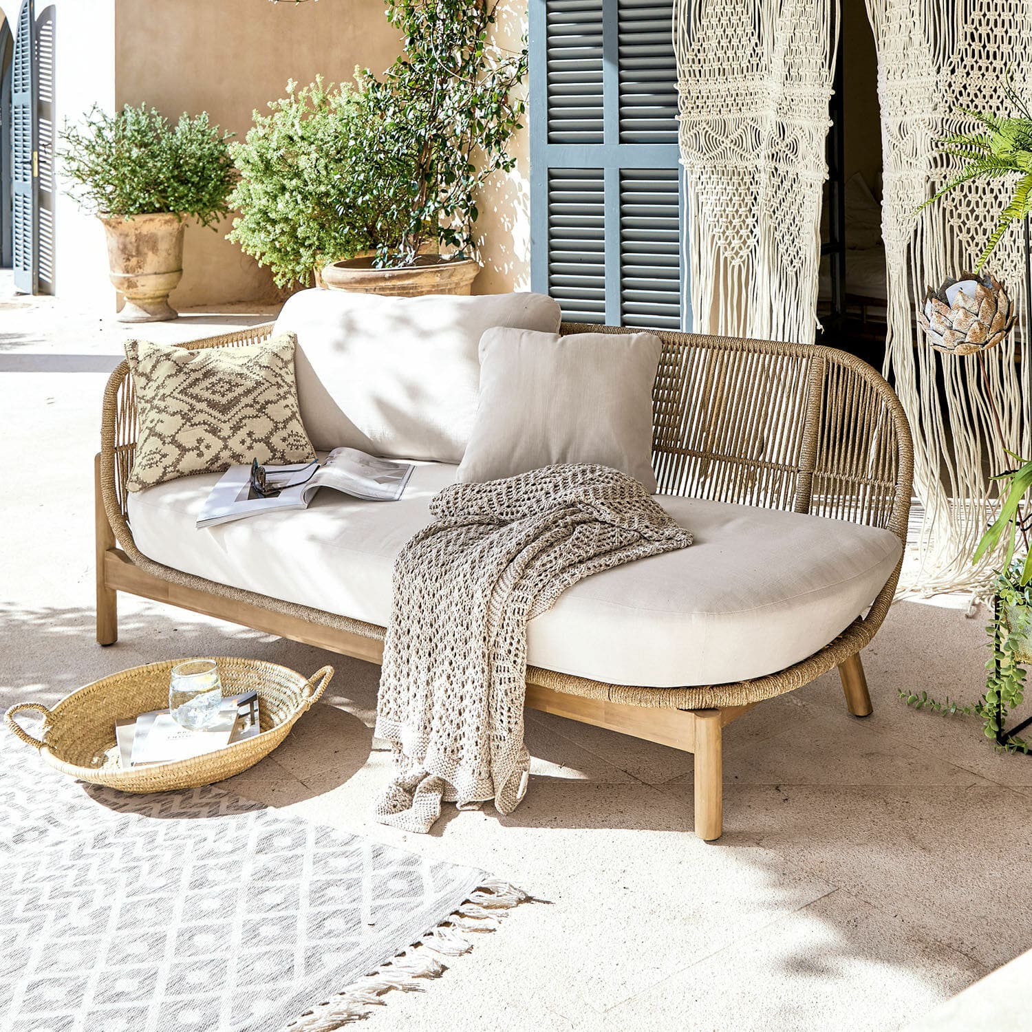 Chaiselongue FSC®-100% Belvidere Rattan- und Holzsofa mit cremefarbenen Kissen und Decke auf heller Terrasse mit Pflanzen