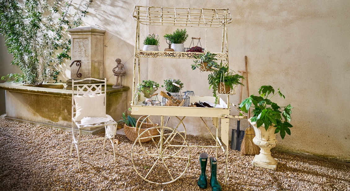 Französischer Gartenstil mit Metallwagen für Pflanzen, grünen Pflanzen und Gartengeräten auf Kiesboden