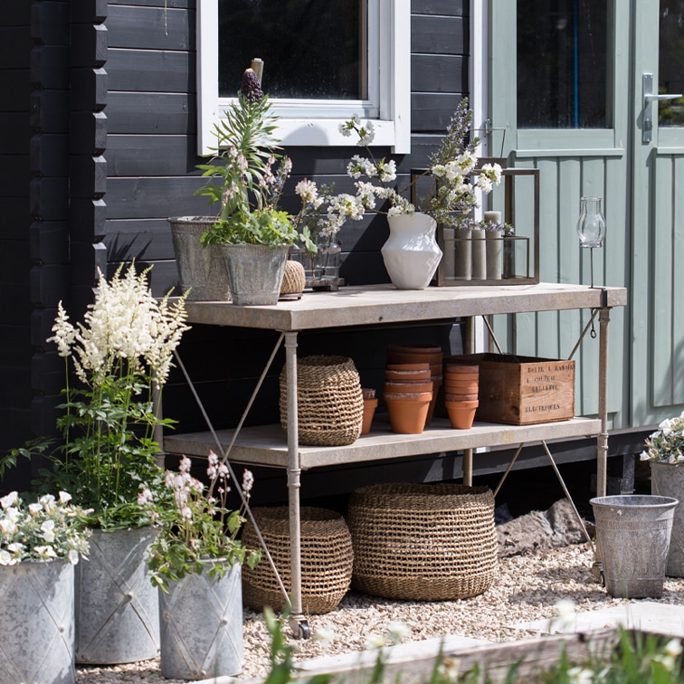 Metall-Gartentisch mit Blumentöpfen, geflochtenen Körben und Blumen vor schwarzer Wand und grüner Türe
