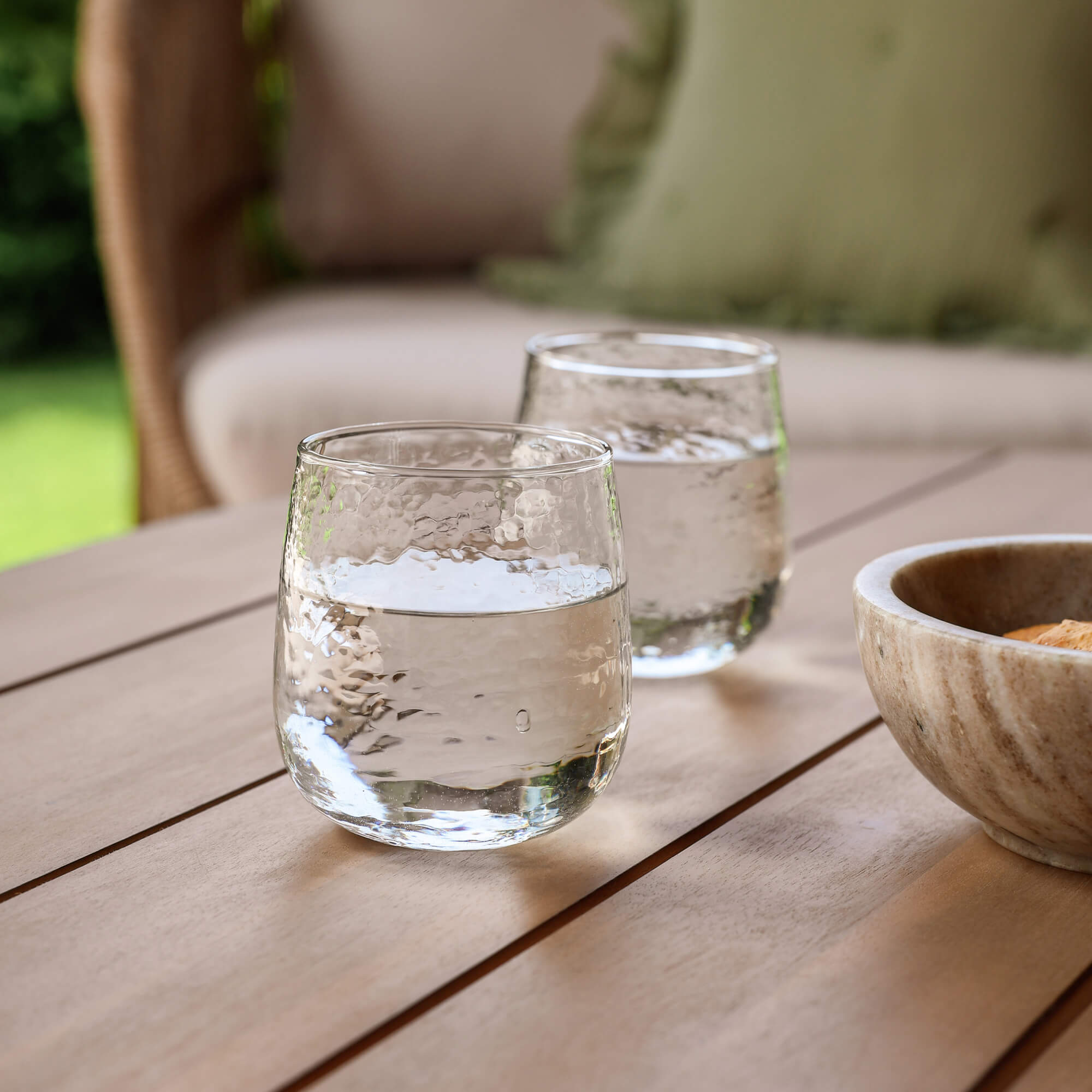 Zwei klare Wassergläser auf einem Holztisch im Garten neben einer Steinschale.