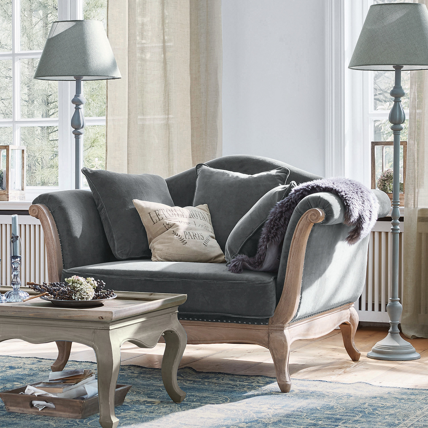 Sessel Sumerset Graues Samtsofa mit Holzrahmen und Kissen in hellem Wohnzimmer mit Stehleuchten