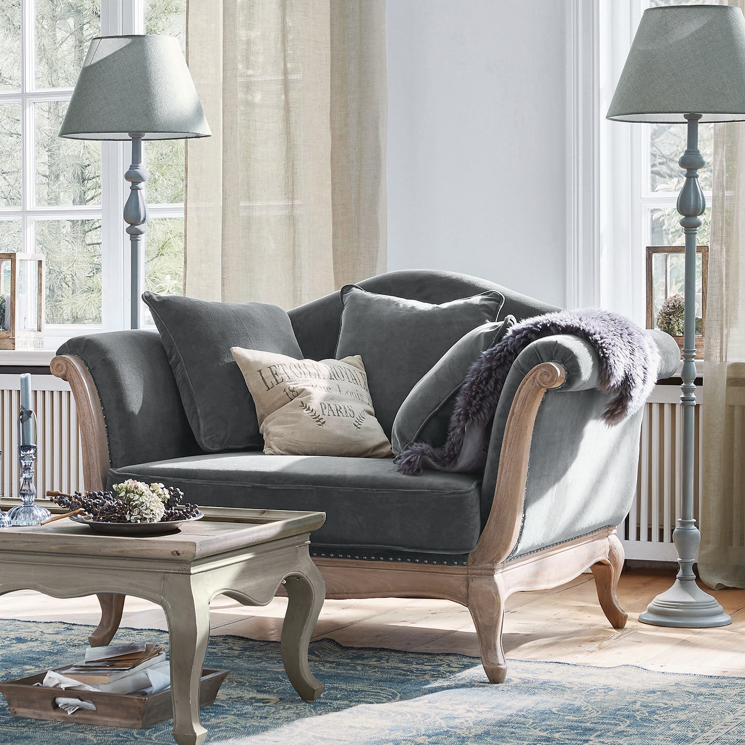 Sessel Sumerset Graues Samtsofa mit Holzrahmen und Kissen in hellem Wohnzimmer mit Stehleuchten