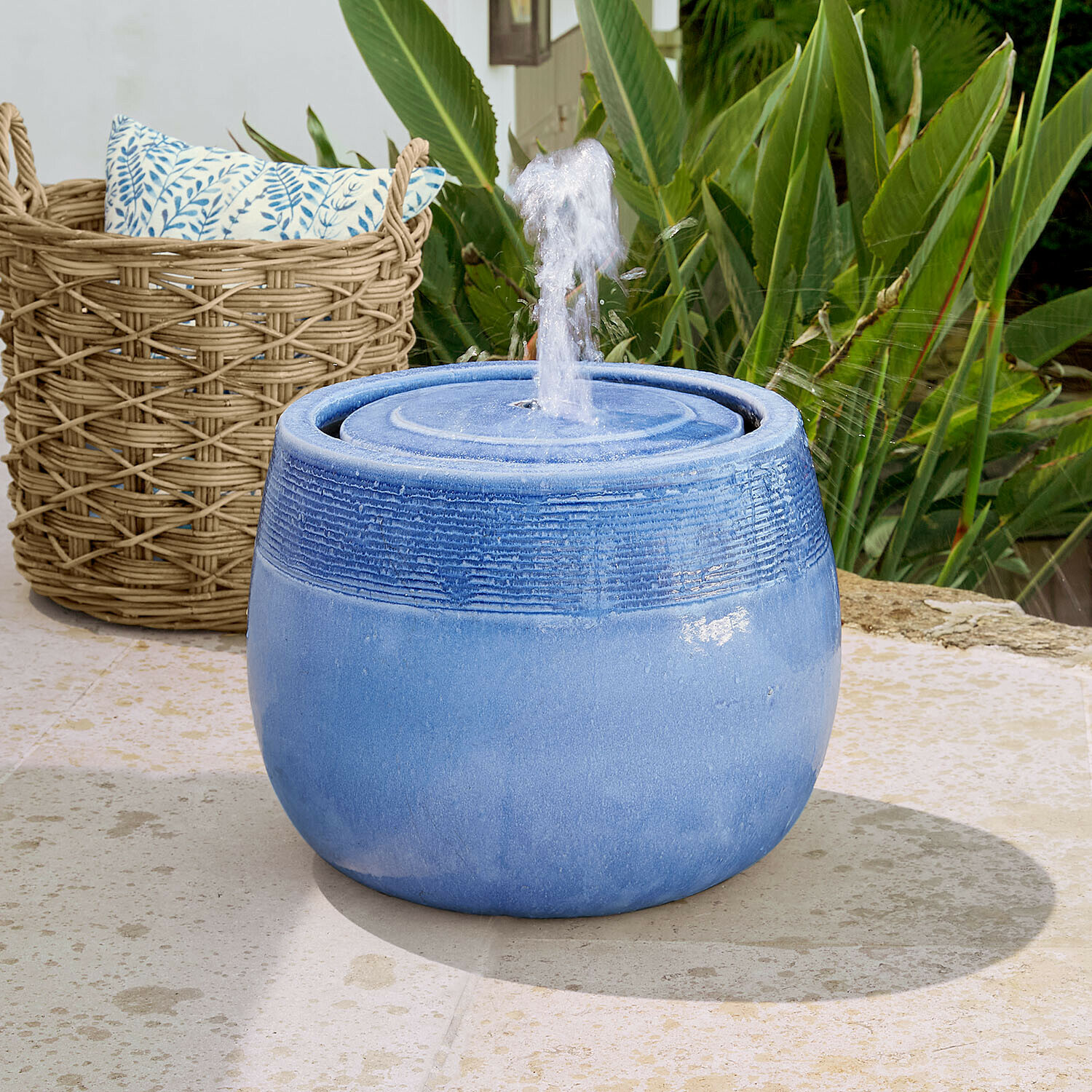 Blaue Keramik-Gartenbrunnen mit spritzendem Wasser vor grünen Pflanzen und Weidenkorb