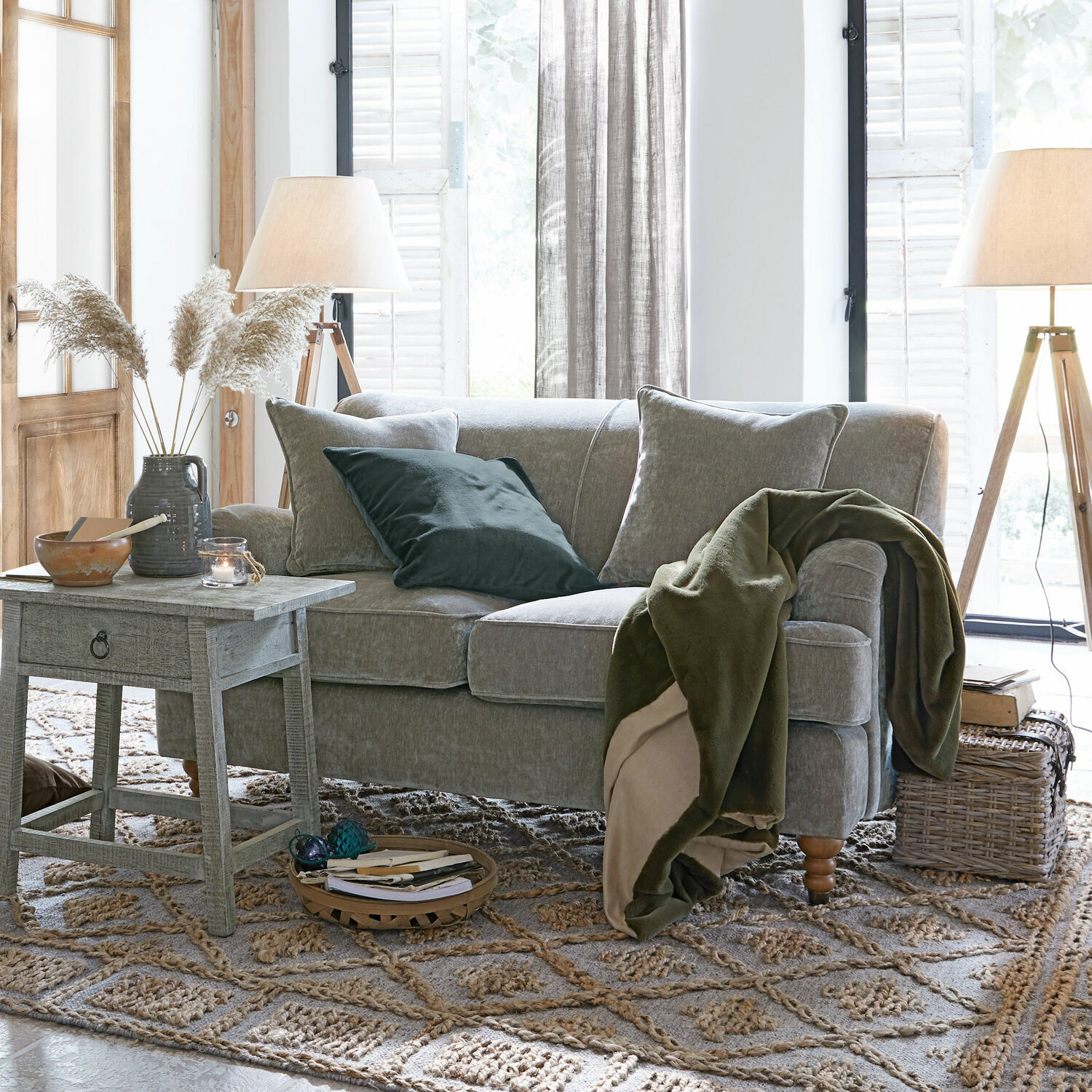 Graues Sofa mit weichen Kissen und grünem Überwurf in hellem Wohnzimmer mit Holzlampen und Teppich.