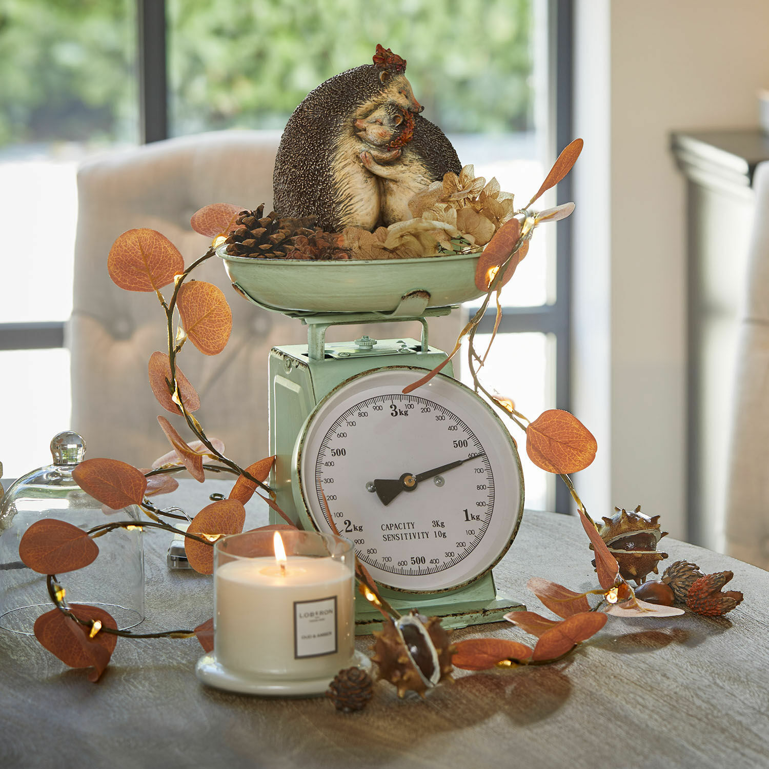 Grüne Metall-Küchenwaage mit Igel-Dekoration und orangefarbenen Blättern auf Tisch mit brennender Kerze