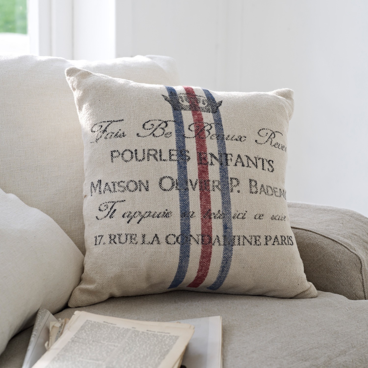 Beige Dekokissen aus Stoff mit Schrift und rot-blauen Streifen auf hellem Sofa im Wohnzimmer