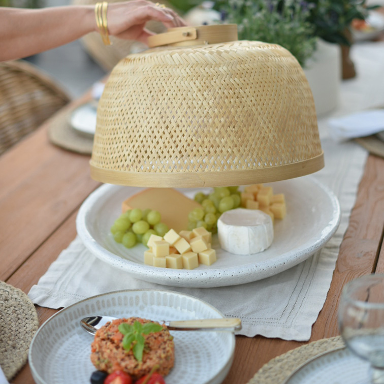 Natürliche Bambus-Abdeckhaube schützt Käse und grüne Trauben auf weissem Teller auf Holztisch