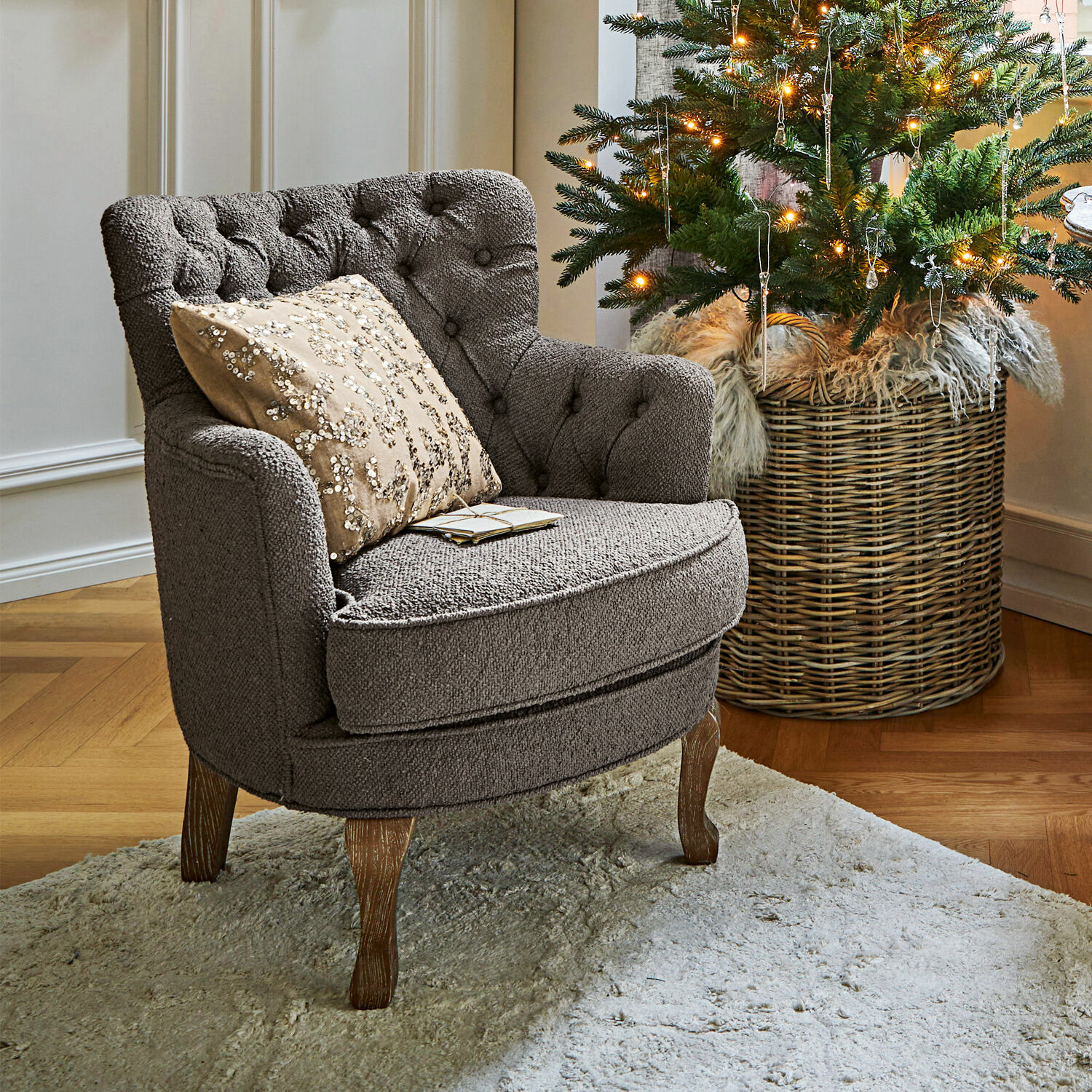 Sessel Calgary Grau gepolsterter Sessel mit Holzbeinen auf hellem Teppich neben geschmücktem Weihnachtsbaum.