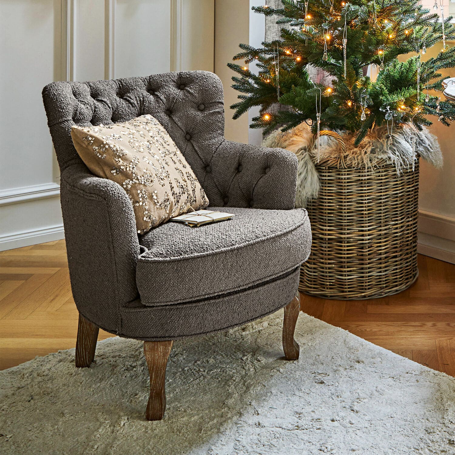 Sessel Calgary Grau gepolsterter Sessel mit Holzbeinen auf hellem Teppich neben geschmücktem Weihnachtsbaum.