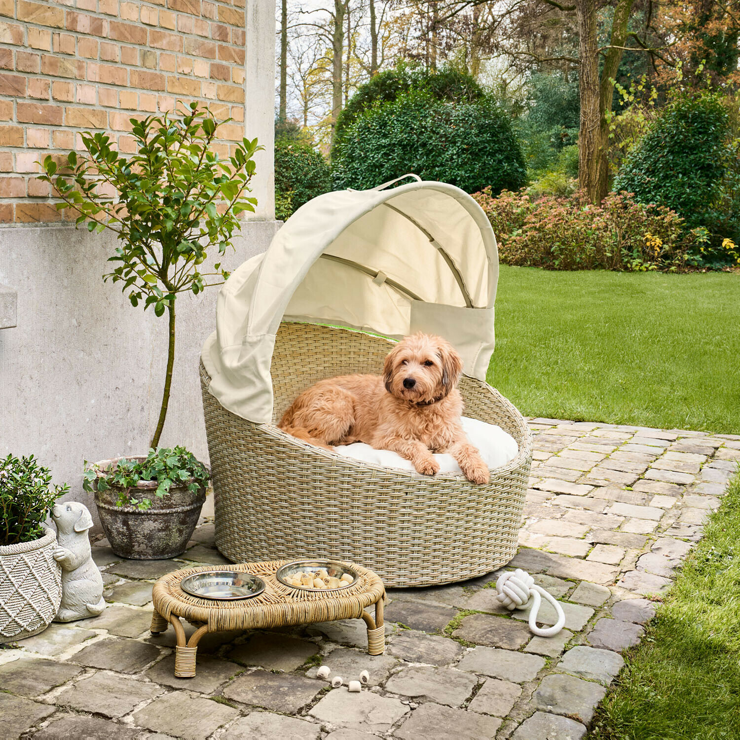 In- & Outdoor-Haustierkorb Brinelza Rundes Hundebett aus beigem Rattan mit cremefarbenem Dach und Kissen auf gepflasterter Terrasse