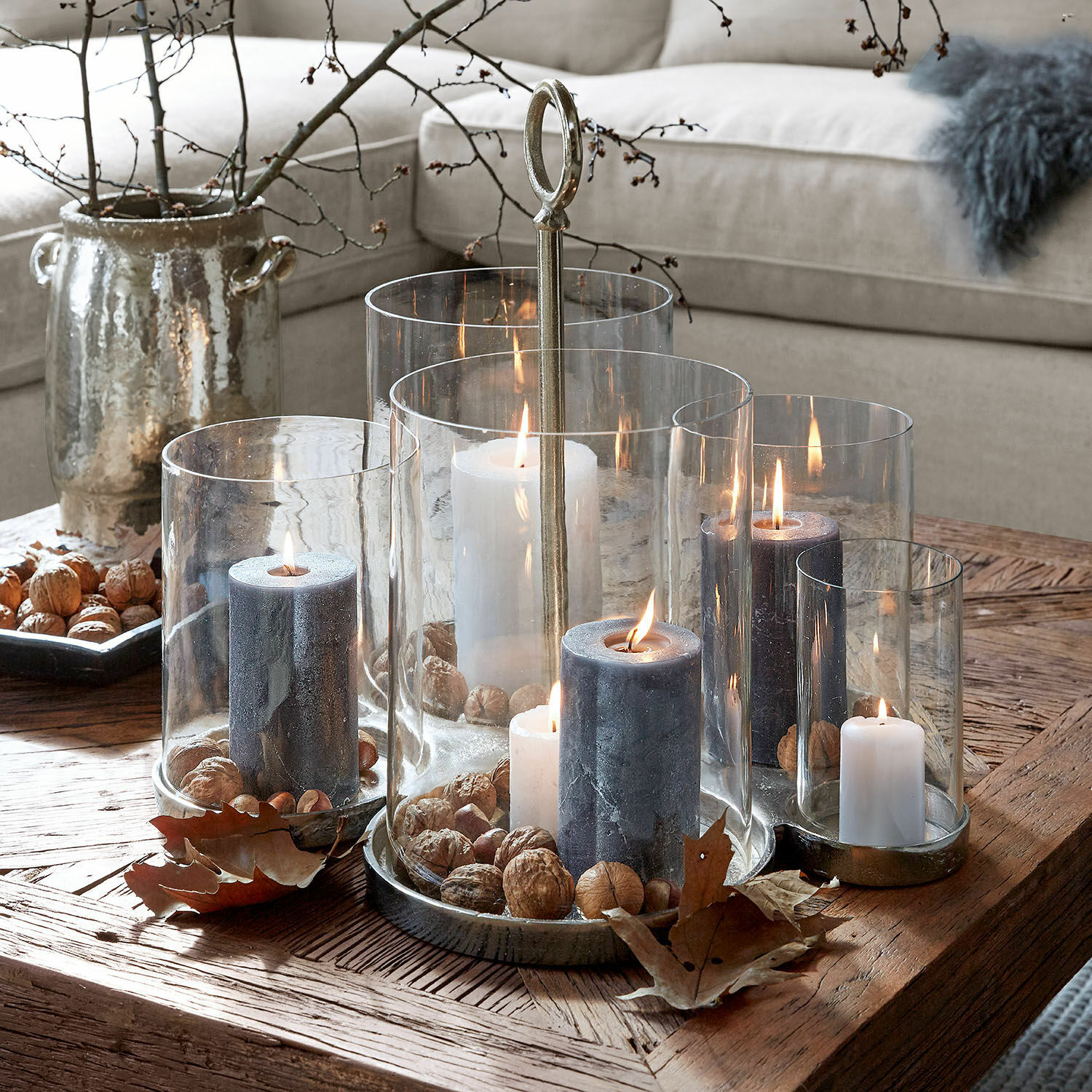 Windlicht Gjaloran Set aus Glas- und Metallkerzenhaltern mit weissen und grauen Kerzen sowie Nüssen auf Holztisch