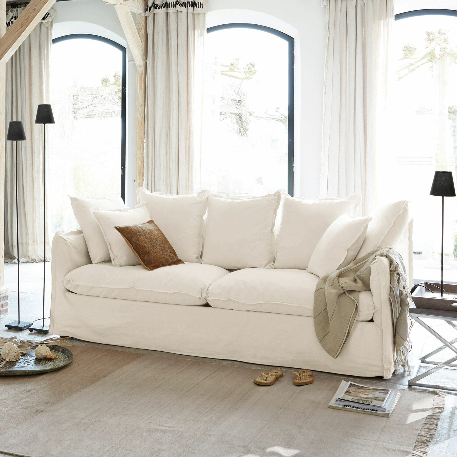 Sofa Tienen Weisses Sofa mit weichen Kissen und braunem Dekokissen in hellem Wohnzimmer mit grossen Fenstern